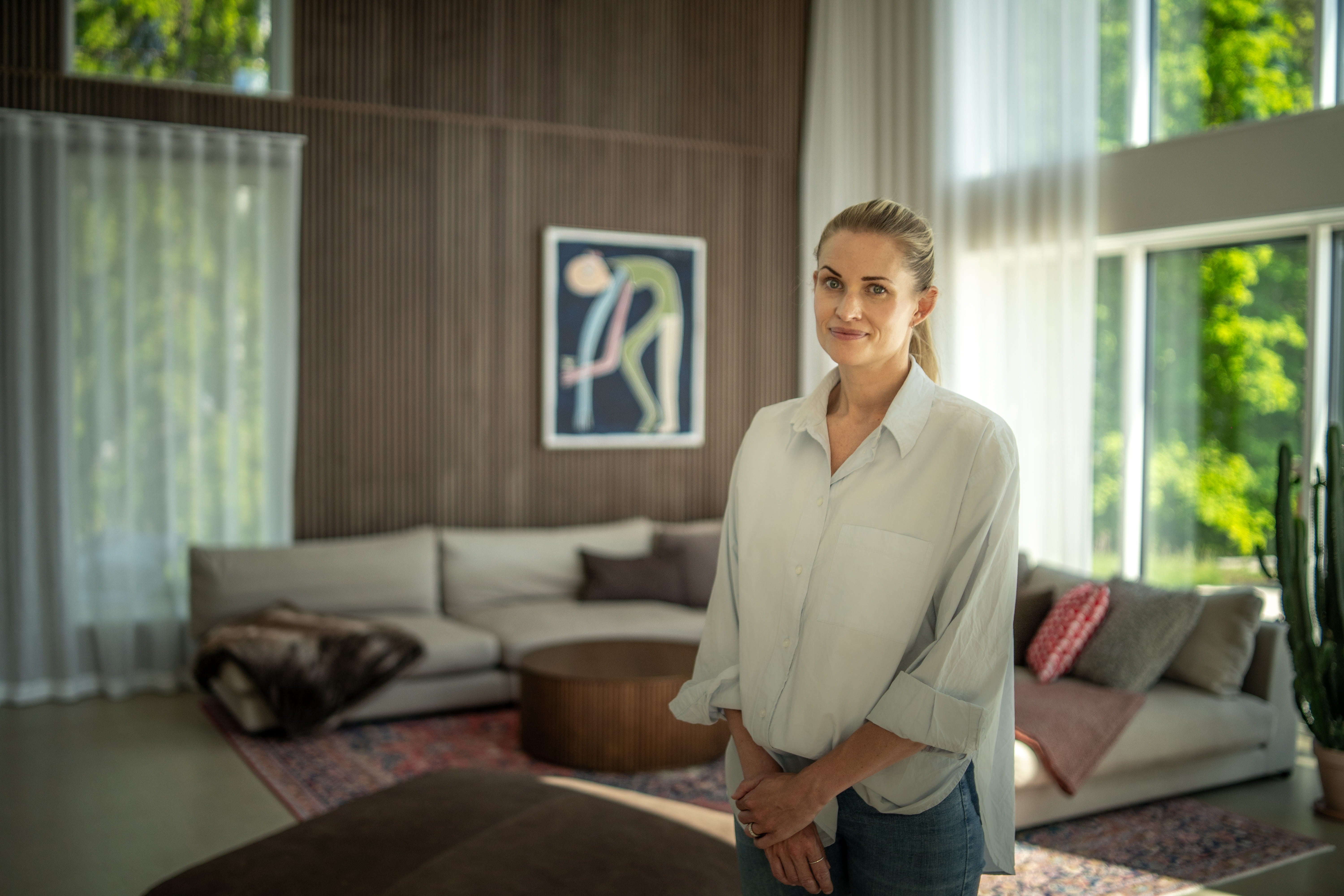 Een vrouw staat in een zonovergoten woonkamer met grote ramen, Scandinavische akoestische houten panelen aan de muur, hedendaagse kunst en neutraal getint minimalistisch meubilair.