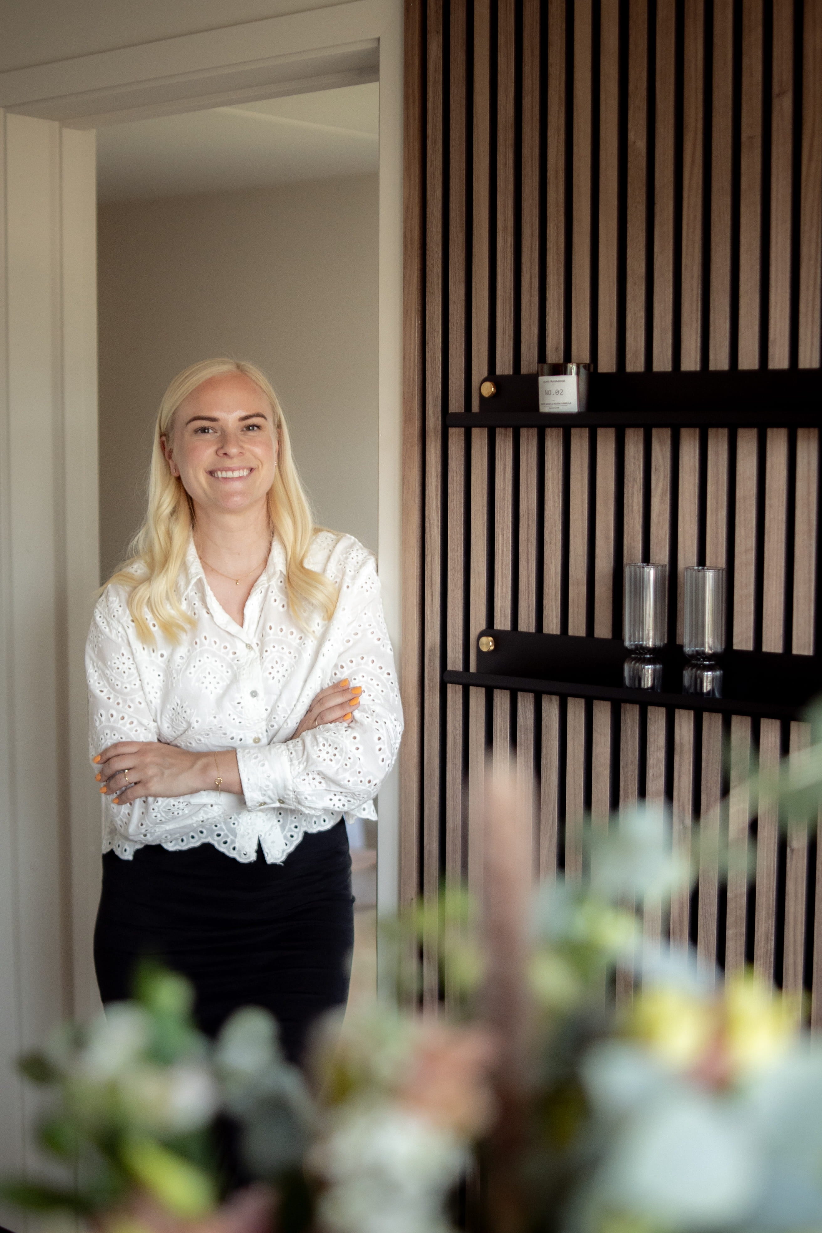 Een vrouw met lang blond haar en een witte blouse staat glimlachend met haar armen over elkaar in een moderne kamer met Scandinavische akoestische houten panelen en strakke planken.