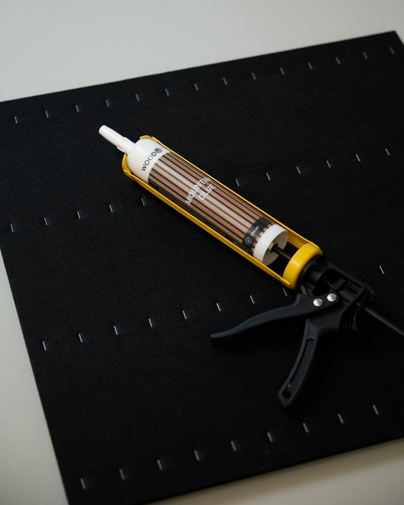 Een geel en black WoodUpp voegkitpistool (290 ml) ligt op een black mat met een raster van witte nietjes, gebruikt voor de installatie van akoestische panelen.