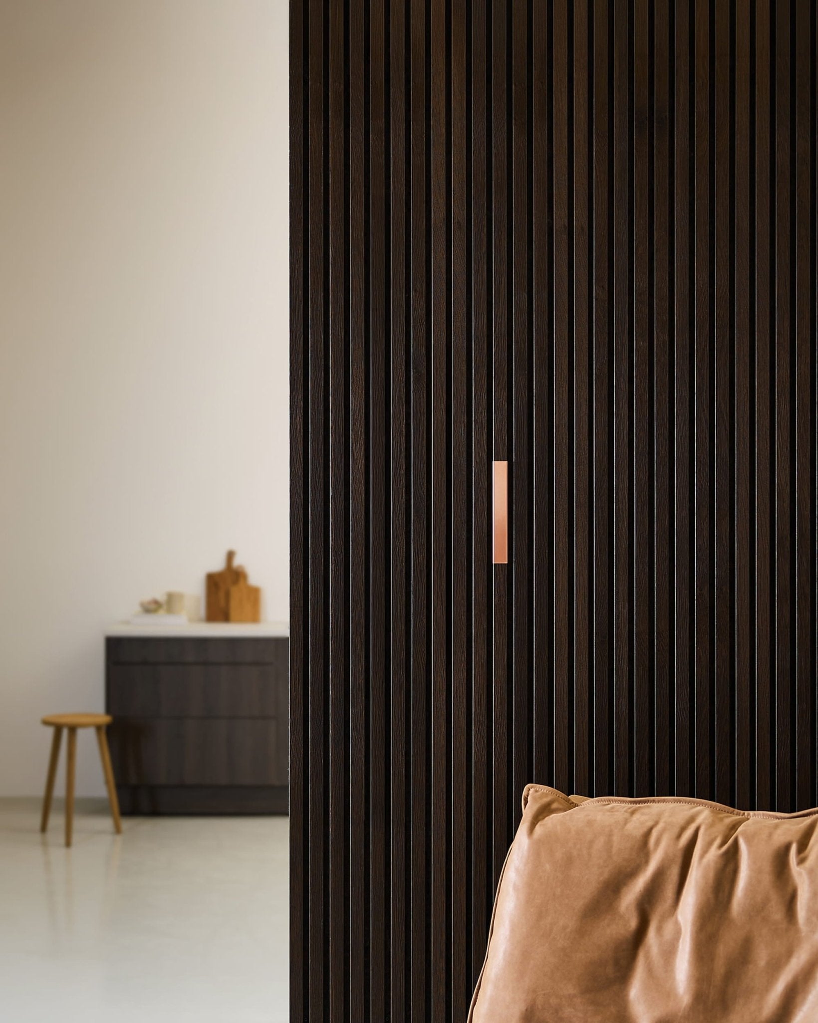 Minimalistisch interieur met een bruine leren stoel, WoodUpp Liva | Cover / Copper akoestische panelen van 20 cm aan de muur, en een kruk bij een aanrecht met snijplanken.