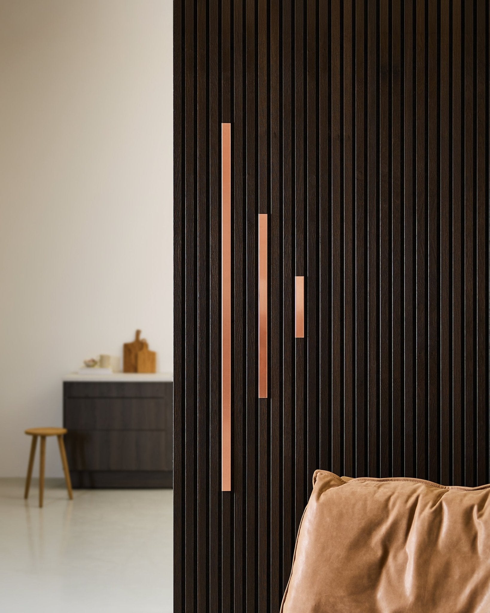 Minimalistische ruimte met een donkere houten akoestische lattenwand met WoodUpp Liva Cover in copper, panelen van 60 cm met copper handgrepen, een lichtbruine leren stoel en een kruk bij de kast.