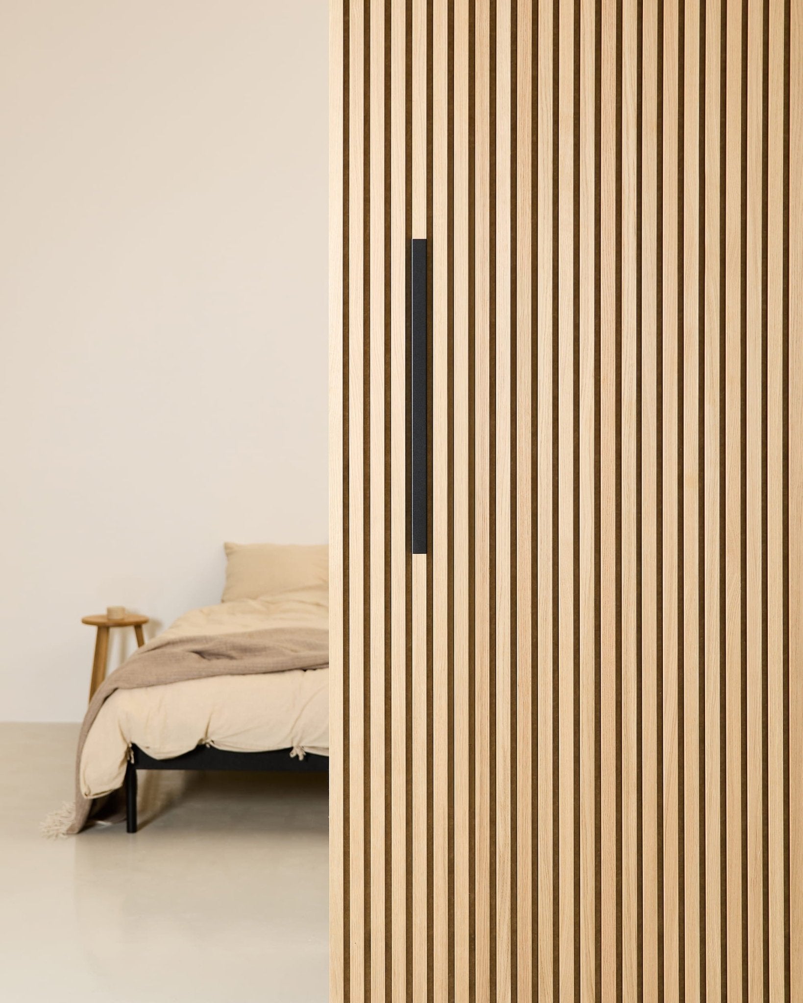 Minimalistische slaapkamer met een beige bed, gedeeltelijk afgeschermd door een 60 cm brede WoodUpp EMBRACE-ruimteverdeler van zwart aluminium met een zwarte handgreep.