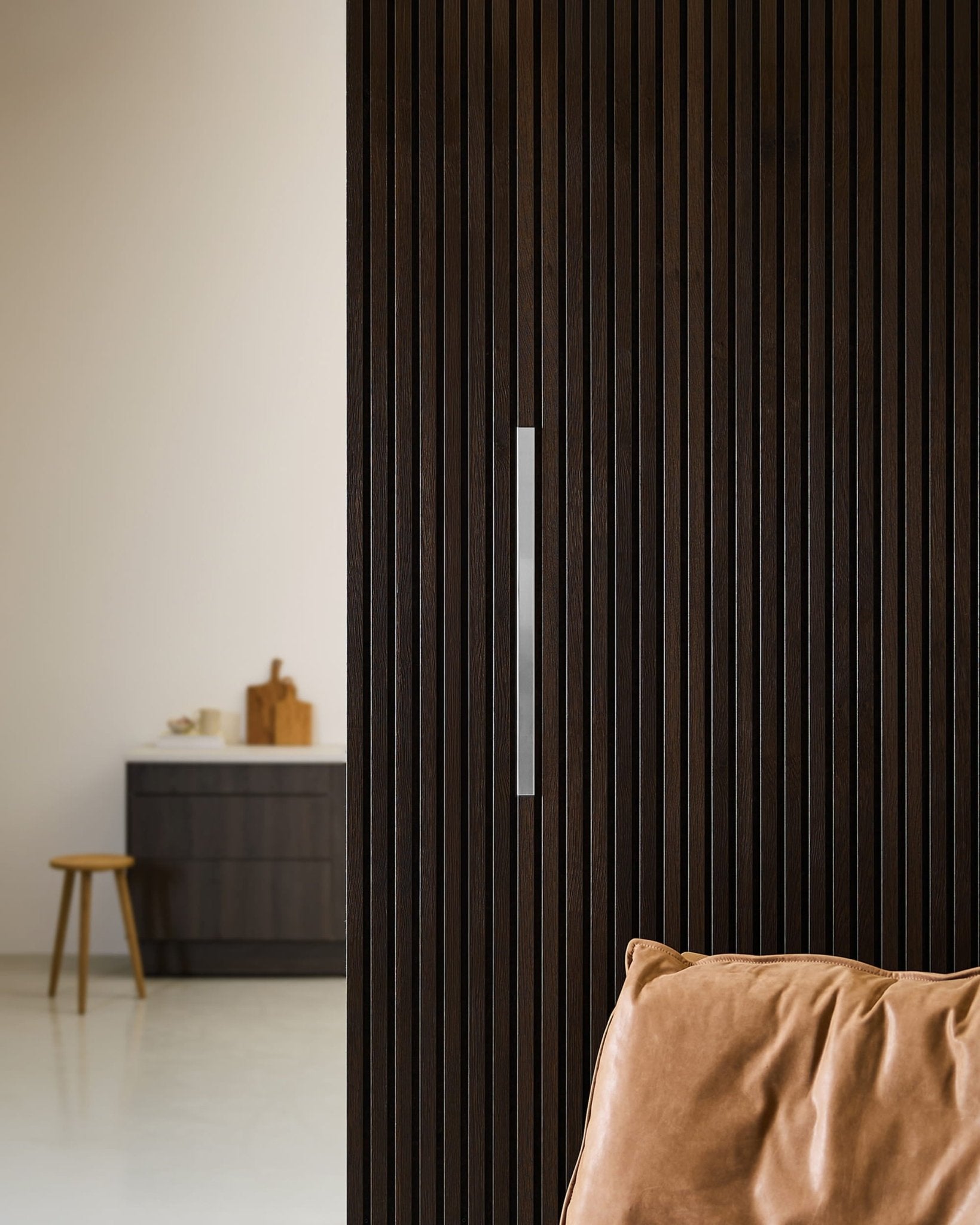 Een moderne Scandinavische ruimte met akoestische panelen van WoodUpp Liva in gepolijst staal, een wand met donkere houten latten, een bruinleren stoel en een kruk naast een keukenkast.