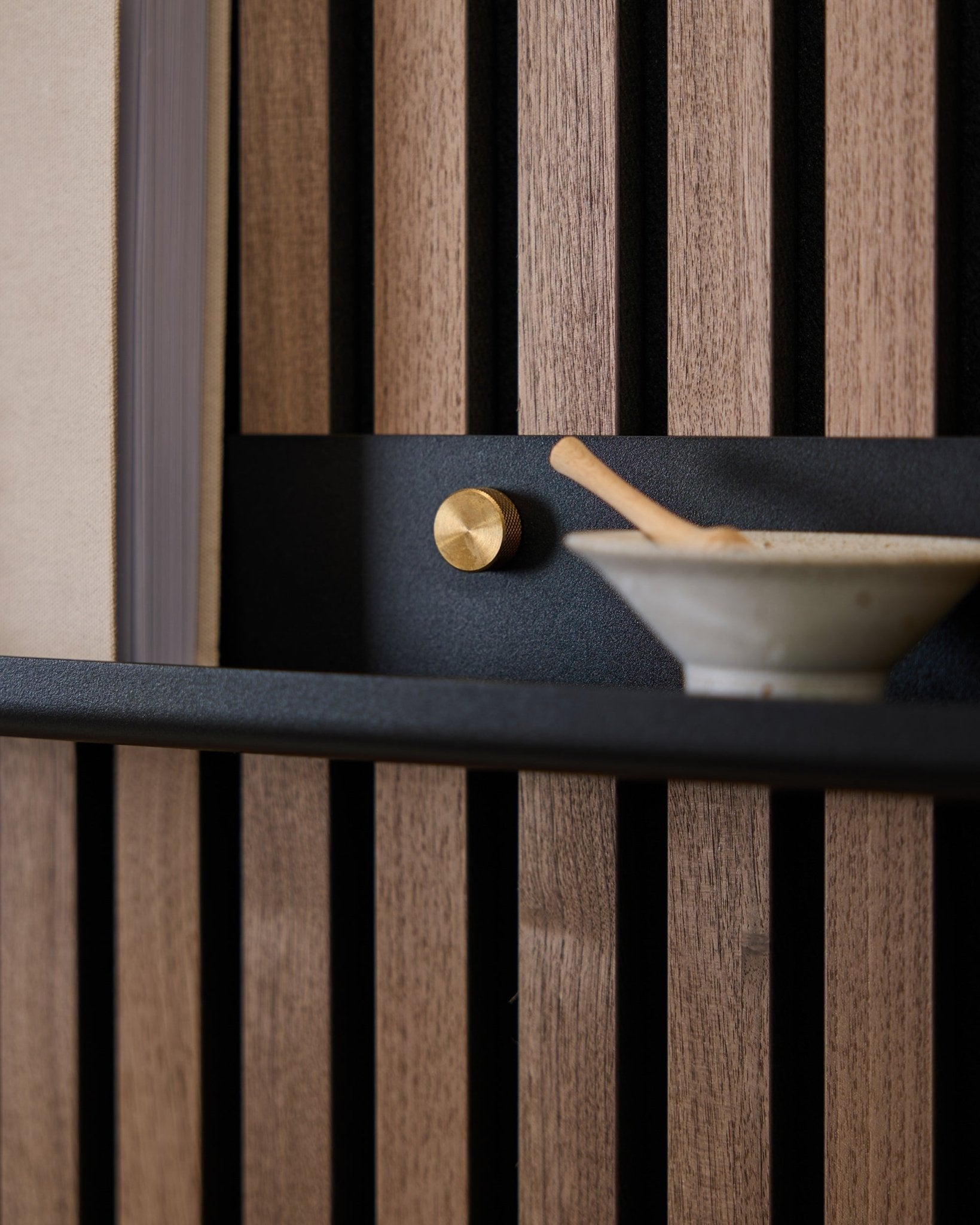 Close-up van een 115,9 cm black WoodUpp Norr Narrow Shelf, gemonteerd op een Scandinavisch akoestisch lattenwandpaneel met brass duimschroeven, een gouden knop en een keramische kom met lepel.