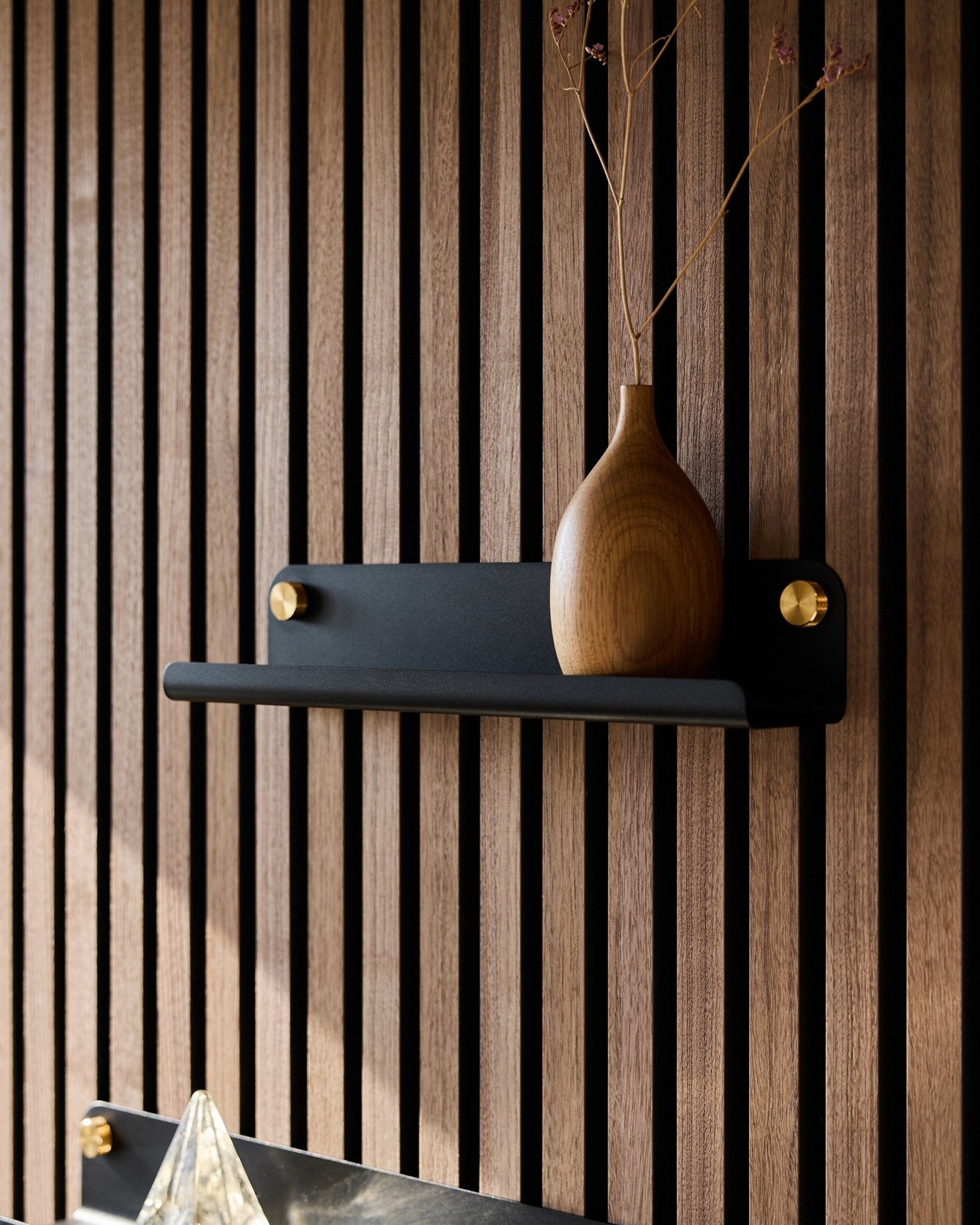 Een houten vaas met gedroogde takken op een black WoodUpp Norr Narrow shelf (35,9 cm), geplaatst voor een vertical akoestisch lattenwandpaneel in Nordic stijl.