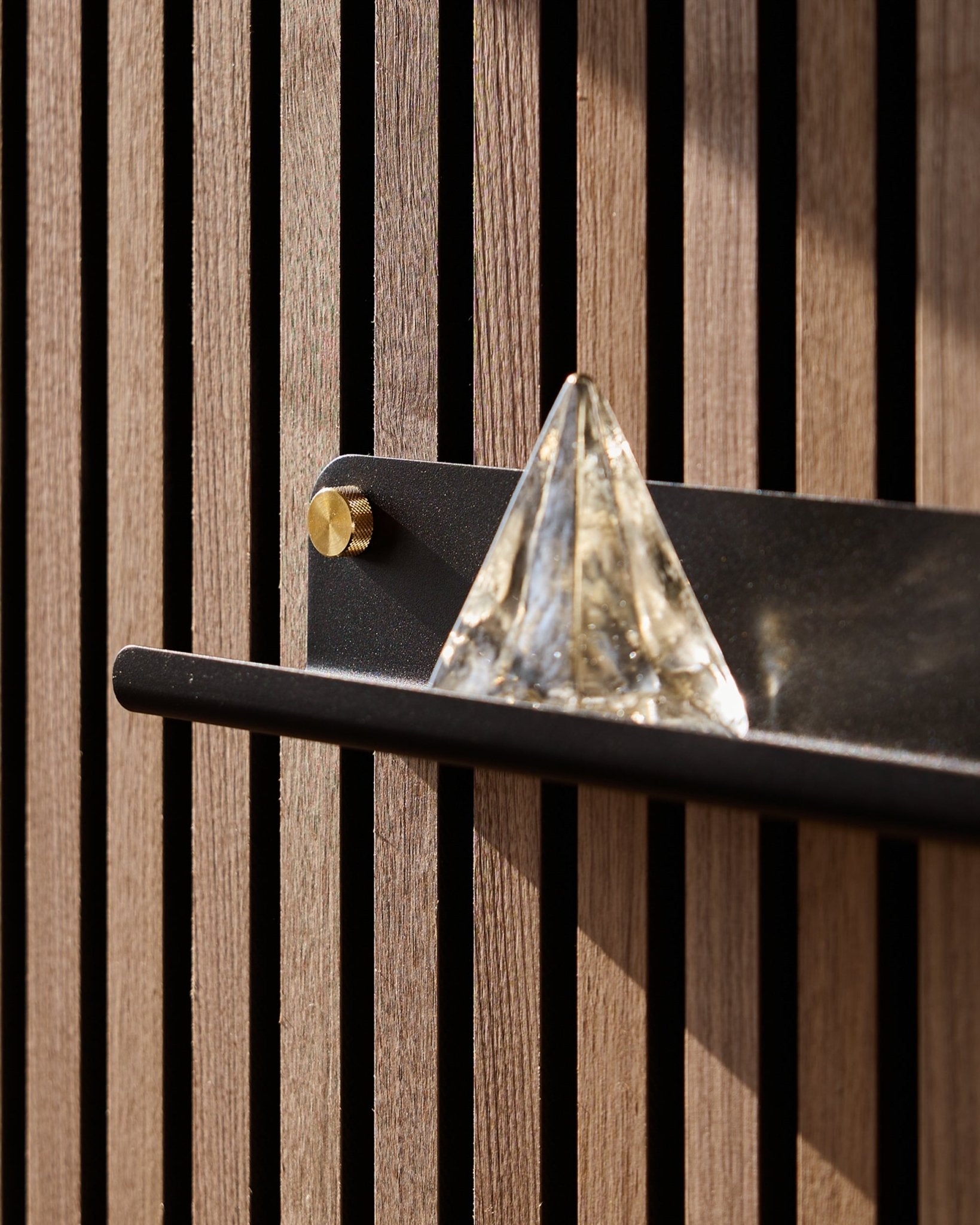 Een glazen piramide staat op de WoodUpp Norr Narrow Shelf in black (51,9 cm), gemonteerd op een vertical Akoestisch paneel en geaccentueerd door zonlicht.