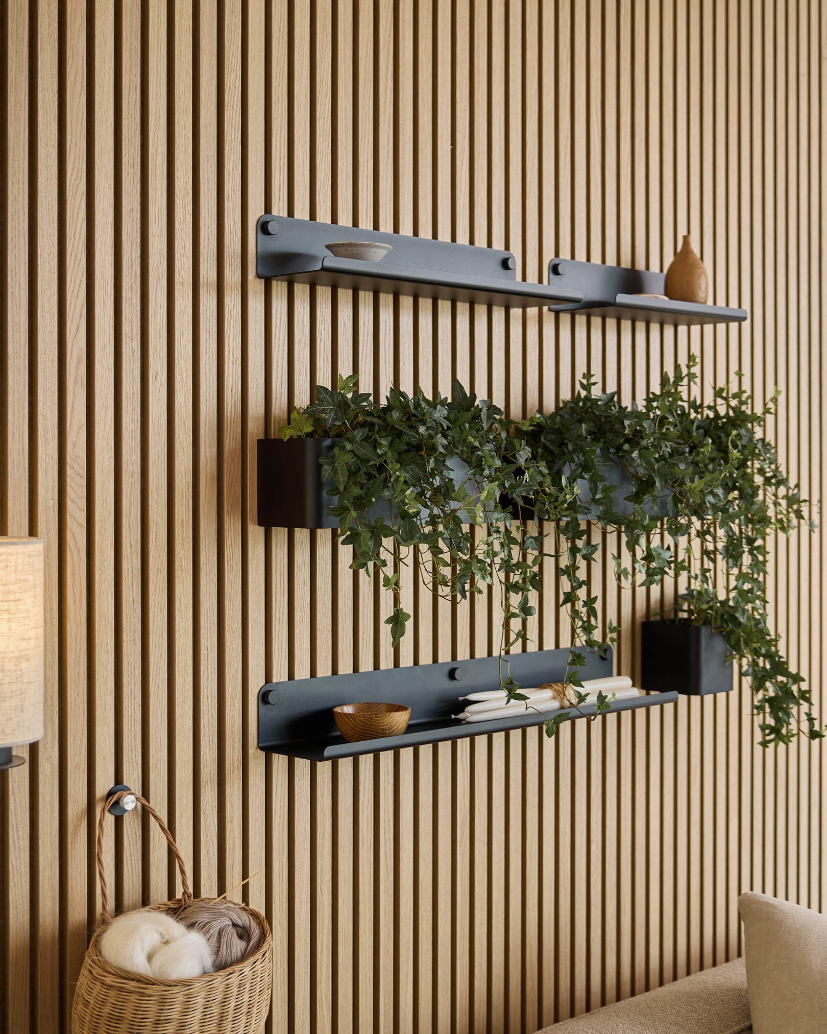 Drie black Norr | Plant Box planken van WoodUpp met decoratie en een plant zijn gemonteerd boven een rieten mand met garen, tegen een groen akoestisch lattenwandpaneel.