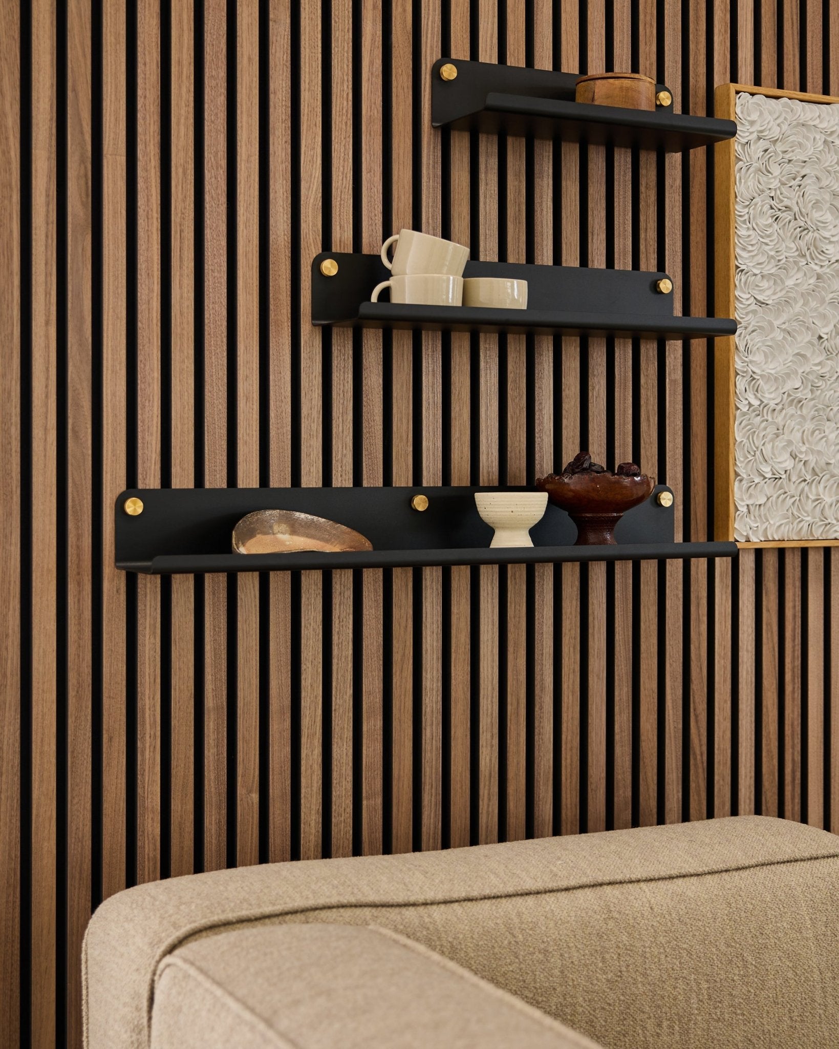Drie black planken, waaronder de WoodUpp Norr Wide Shelf (75,9 cm), tonen decoratie op een houten lattenwand in Akoestisch paneel boven een beige bank; een deel van het kunstwerk is zichtbaar.