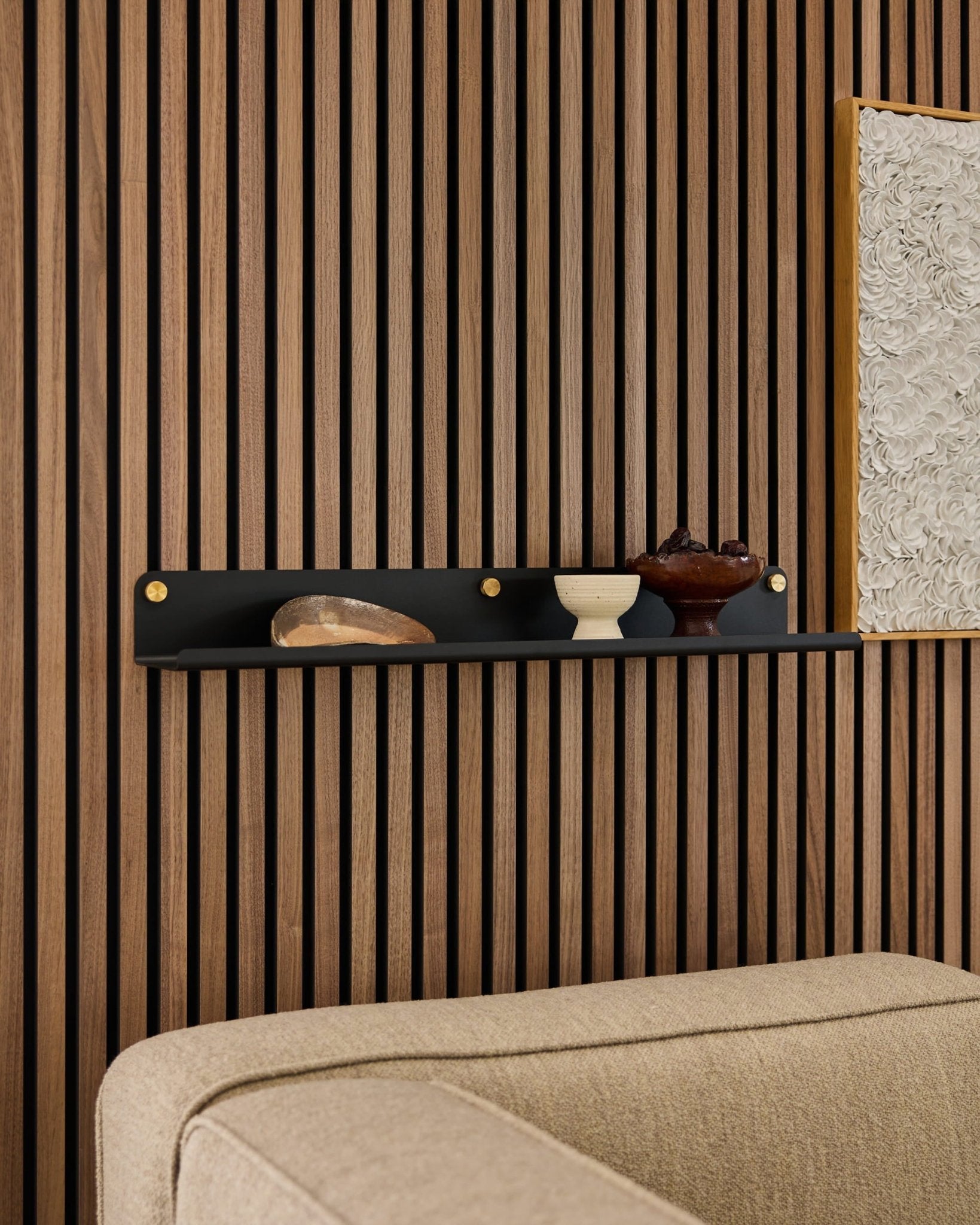 Zwarte WoodUpp Norr brede plank (75,9 cm) met decor boven een beige bank, bevestigd aan een modern houten akoestisch lamellenpaneel.