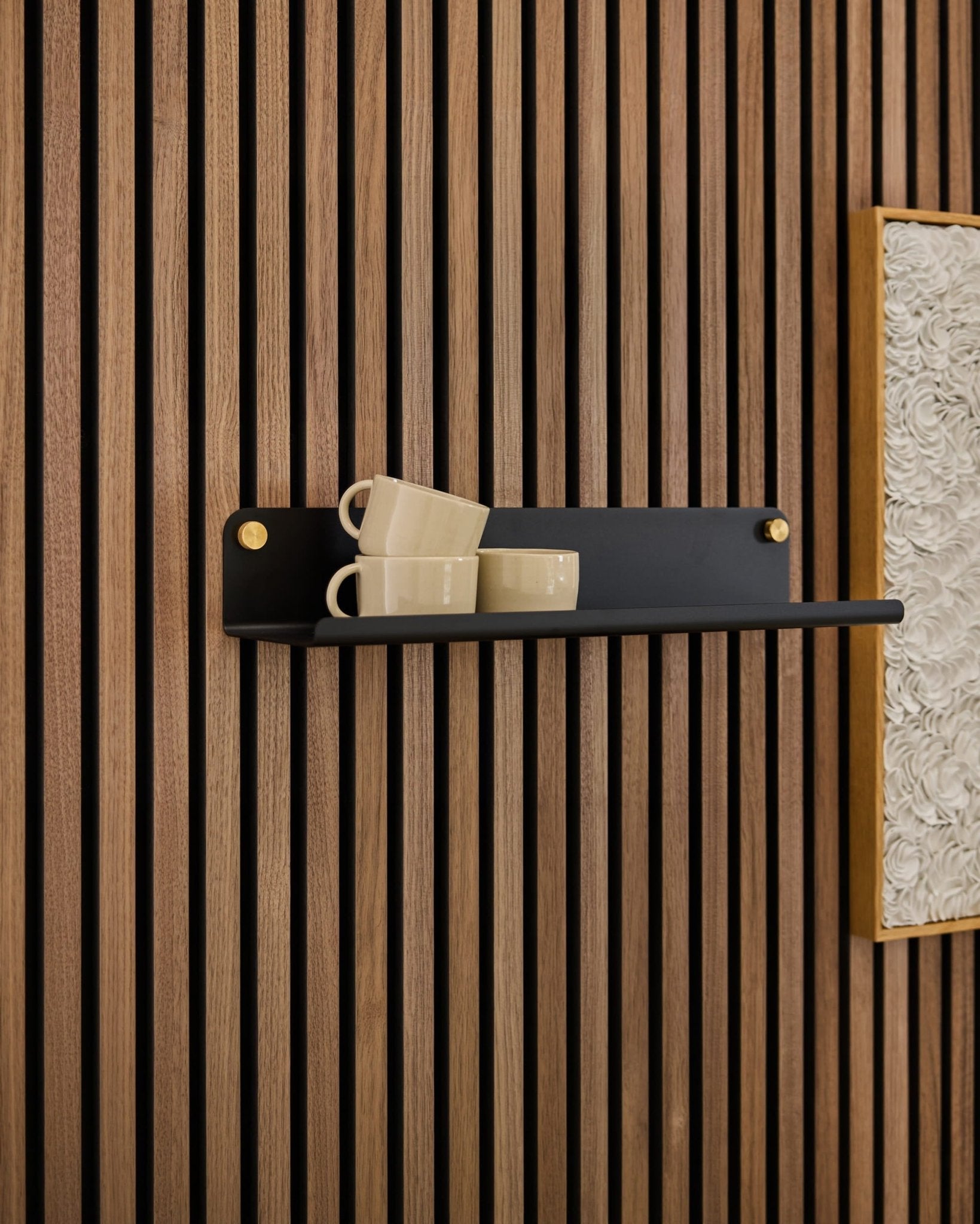 Op een zwarte Norr Wide Shelf van WoodUpp staan drie beige mokken, bevestigd aan een Akupanel-akoestisch paneel naast een ingelijste, witte kunstprint met structuur.
