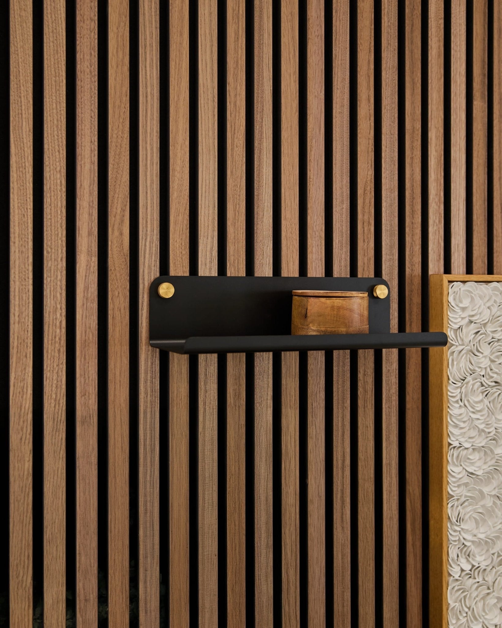 Een klein houten kistje op een WoodUpp Norr Wide-plank (zwart, 35,9 cm) die tegen verticale Akupanel-akoestische houten latten is bevestigd, waarbij een wit paneel met structuur zichtbaar is.