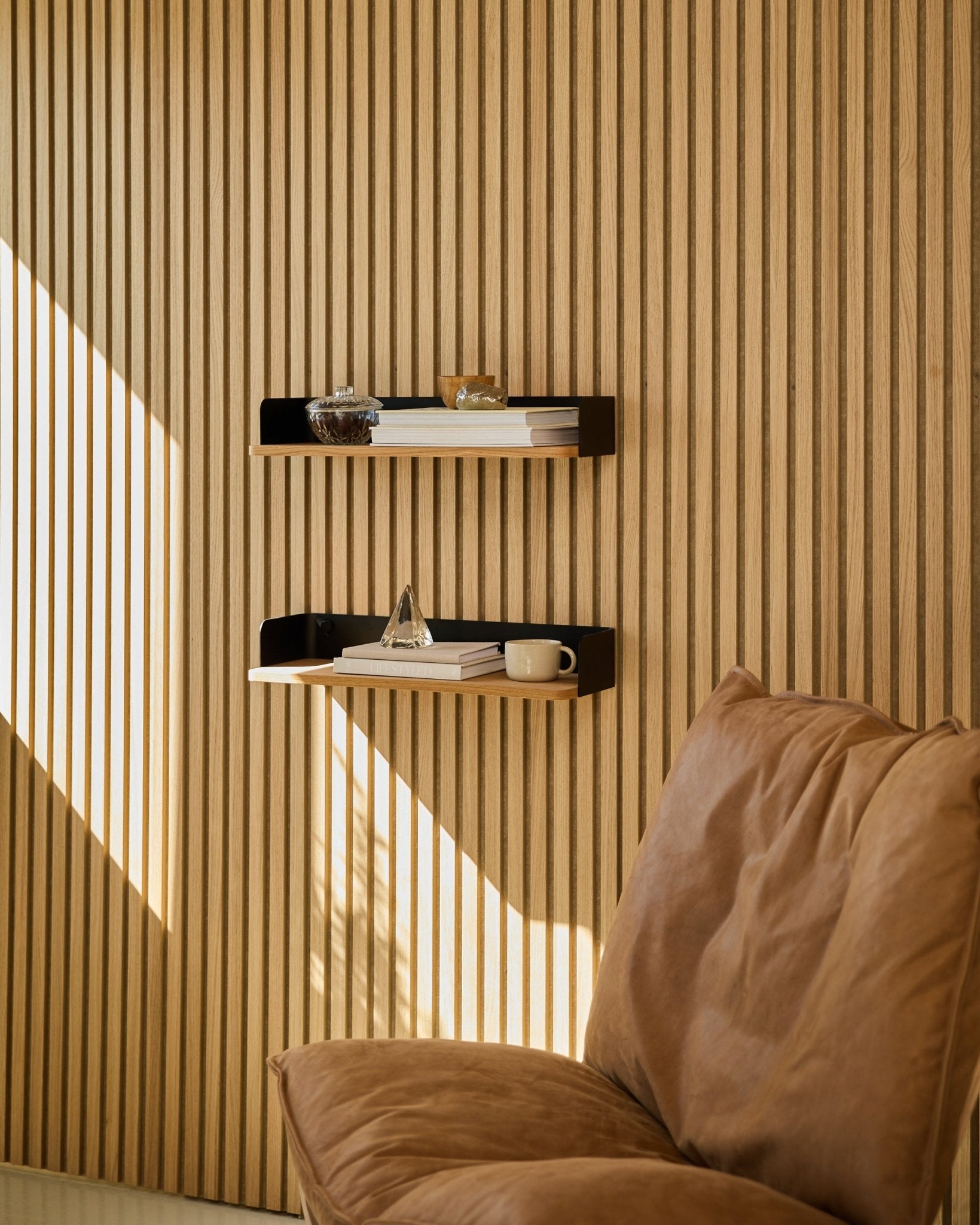 Brown stoel naast de WoodUpp Linn boekenkast in Natural Oak, met boeken en decoratie in natuurlijk licht geplaatst bij een Scandinavische houten lattenwand Akoestisch paneel.