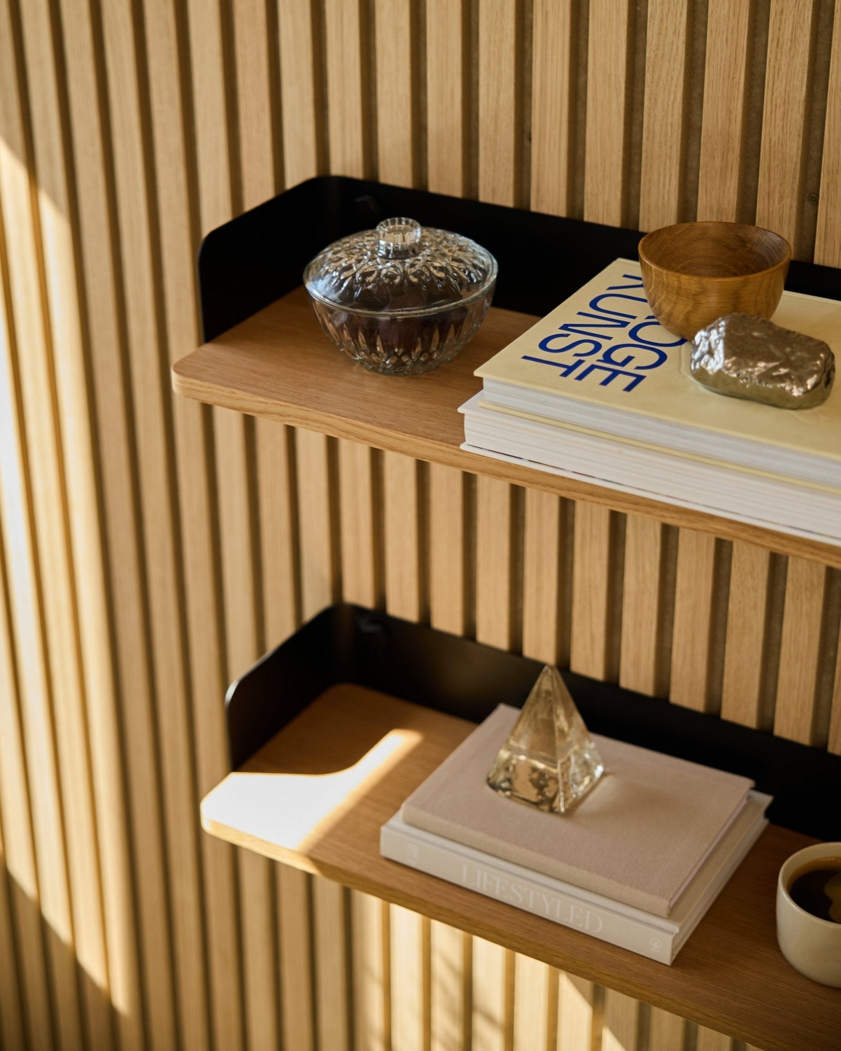 WoodUpp Linn boekenkast in Natural Oak (55,9 cm) met een glazen pot, kopje, gouden piramide en boeken tegen Akupanel akoestische houten panelen in het zonlicht.