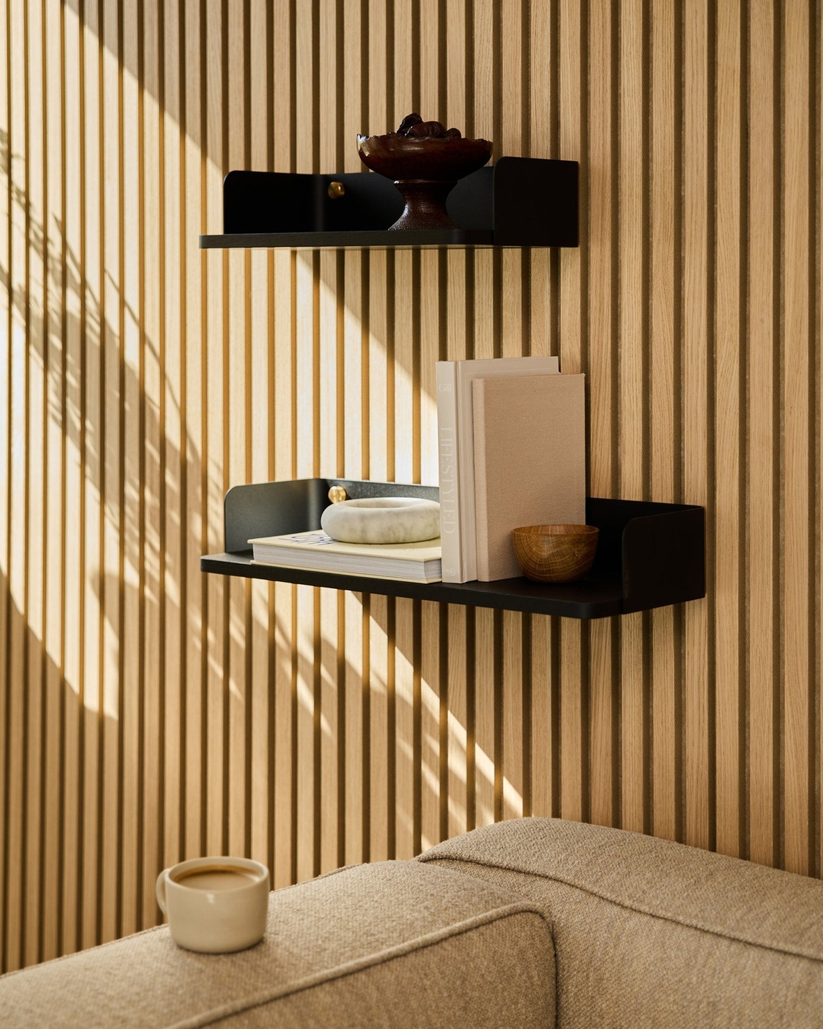 Een black oak WoodUpp boekenkast (55,9 cm) toont boeken, decoratie en een schaal op een wand met akoestische houten panelen; een beige bank en mok op de voorgrond.