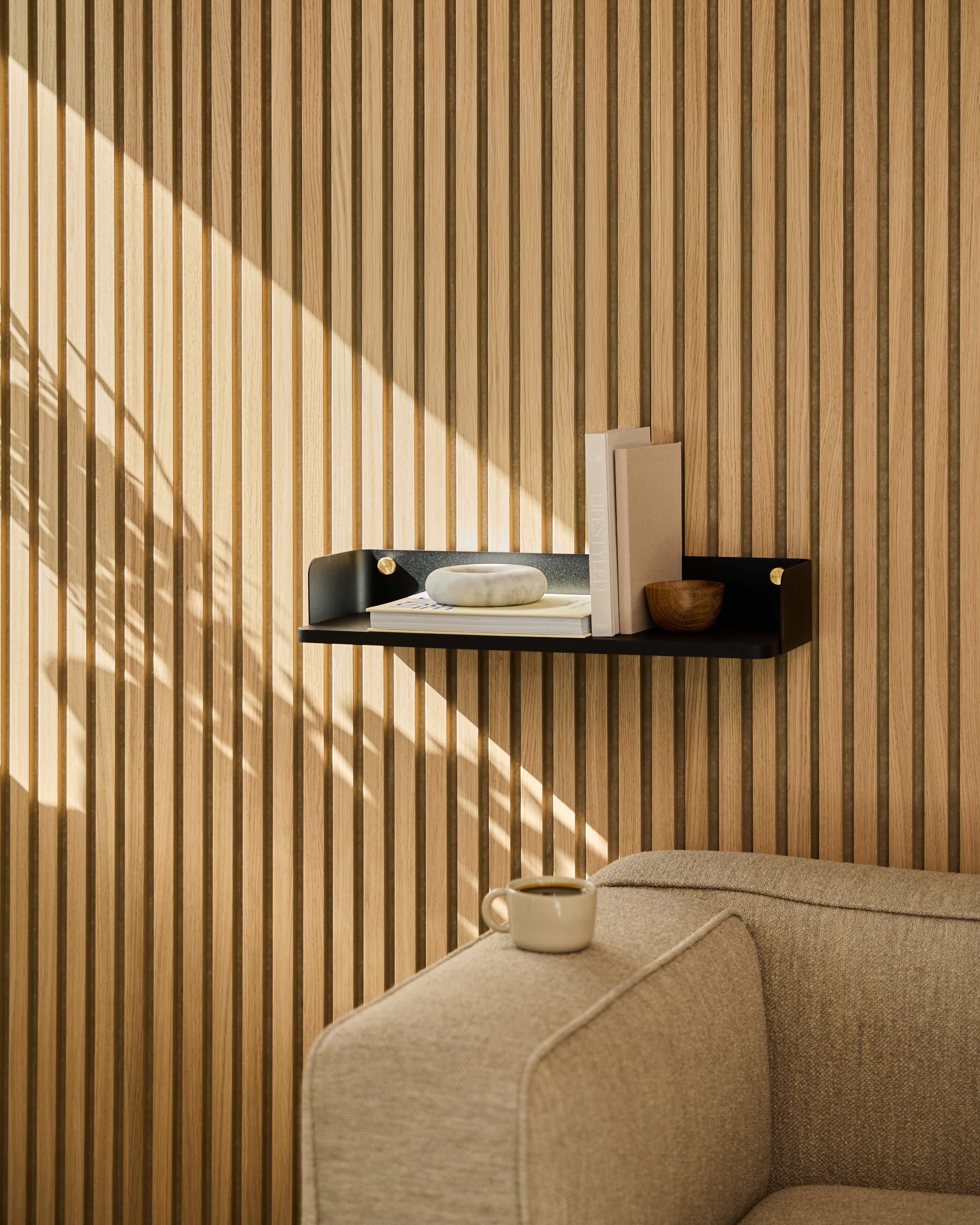 Een Linn | boekenplank / Black Oak van WoodUpp siert de muur boven een beige bank. Het zonlicht werpt schaduwen op de muur met gestreepte akoestische houten panelen.