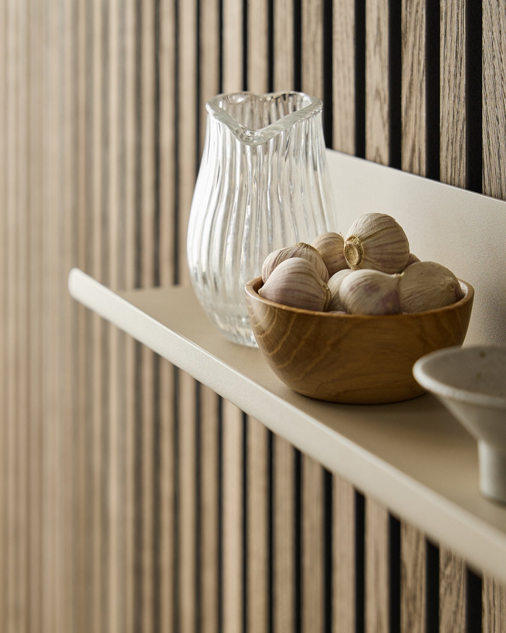 Een houten schaal met knoflook en een glazen karaf op de Norr brede plank van WoodUpp, gemonteerd tegen een Akupanel Akoestisch paneel met vertical houten latten.