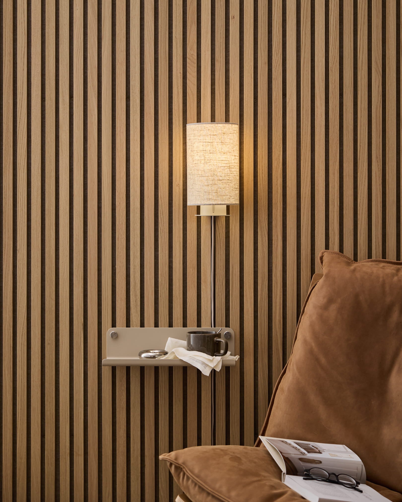 Een beige WoodUpp Feya wandlamp van linnen-katoen, gemonteerd boven een comfortabele fauteuil en bijzettafel met boek en kopje, tegen een Noordse houten lattenwand in Akoestisch paneel.