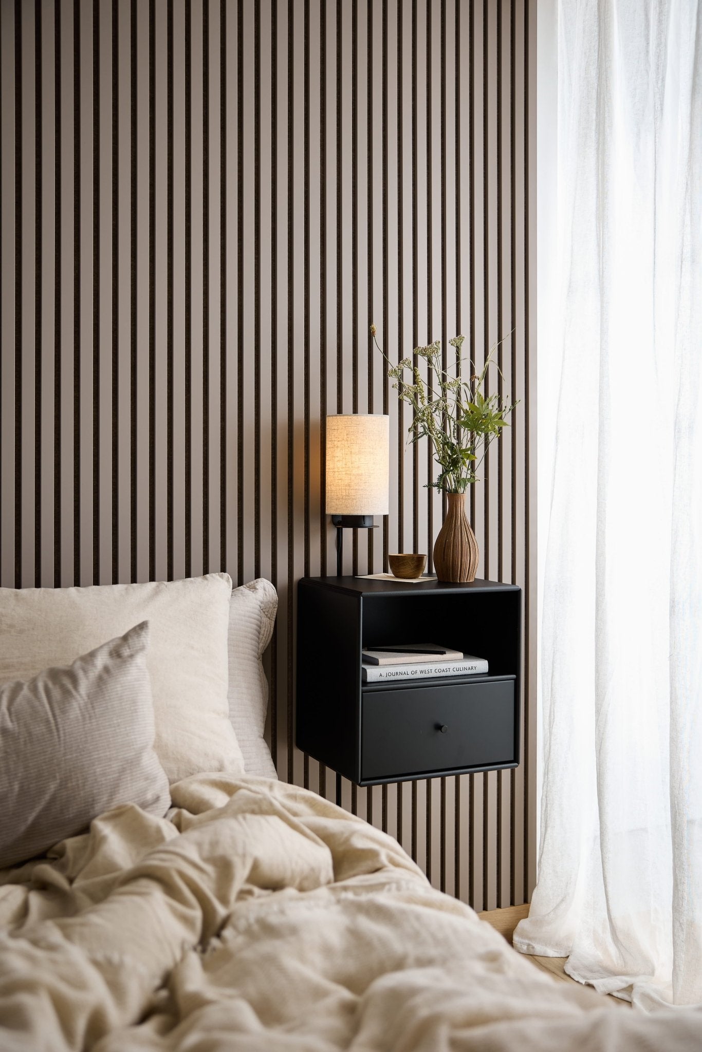 Een bed met beige beddengoed, een zwarte WoodUpp Arka-boekenkast met lade, een lamp en een vaas bij een raam met witte, doorschijnende gordijnen.