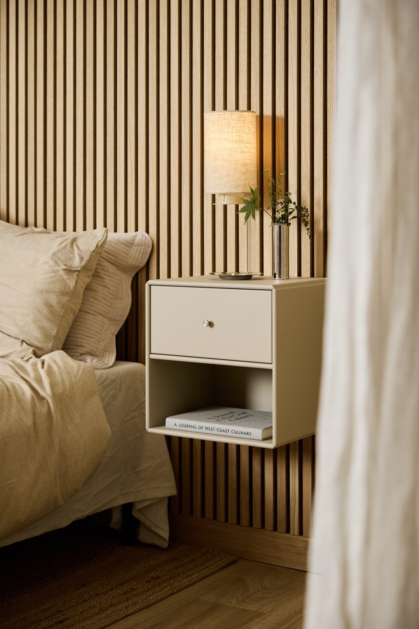 Op een beige WoodUpp Arka-boekenkast met lades naast het bed staan een lamp, boeken en een vaas, met op de achtergrond een WoodUpp Akupanel akoestisch houten lamellenpaneel.