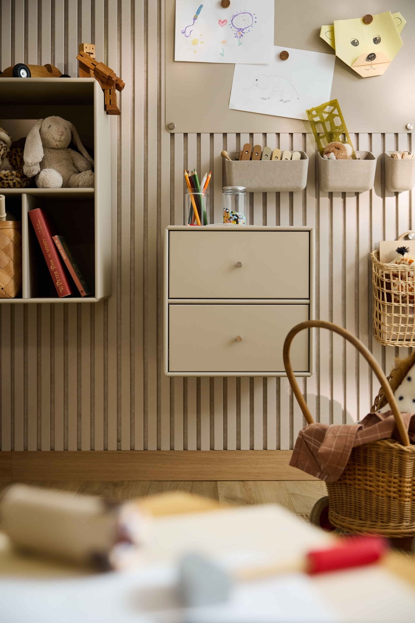 Kinderkamer met gestreept behang, speelgoed, boeken, manden en een beige WoodUpp Arka-boekenkast met lades voor gesloten opbergruimte.
