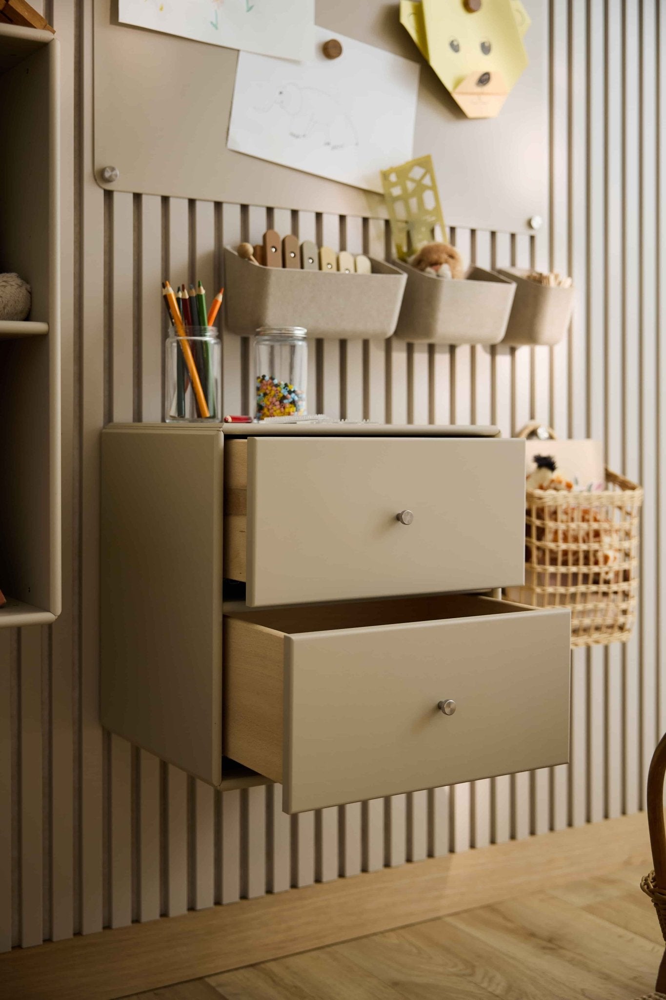 Twee beige Arka-boekenkastladen van WoodUpp, die gedeeltelijk openstaan, bieden ruimte aan knutselspullen en speelgoed; daarnaast staan er gesloten opbergkasten en manden.