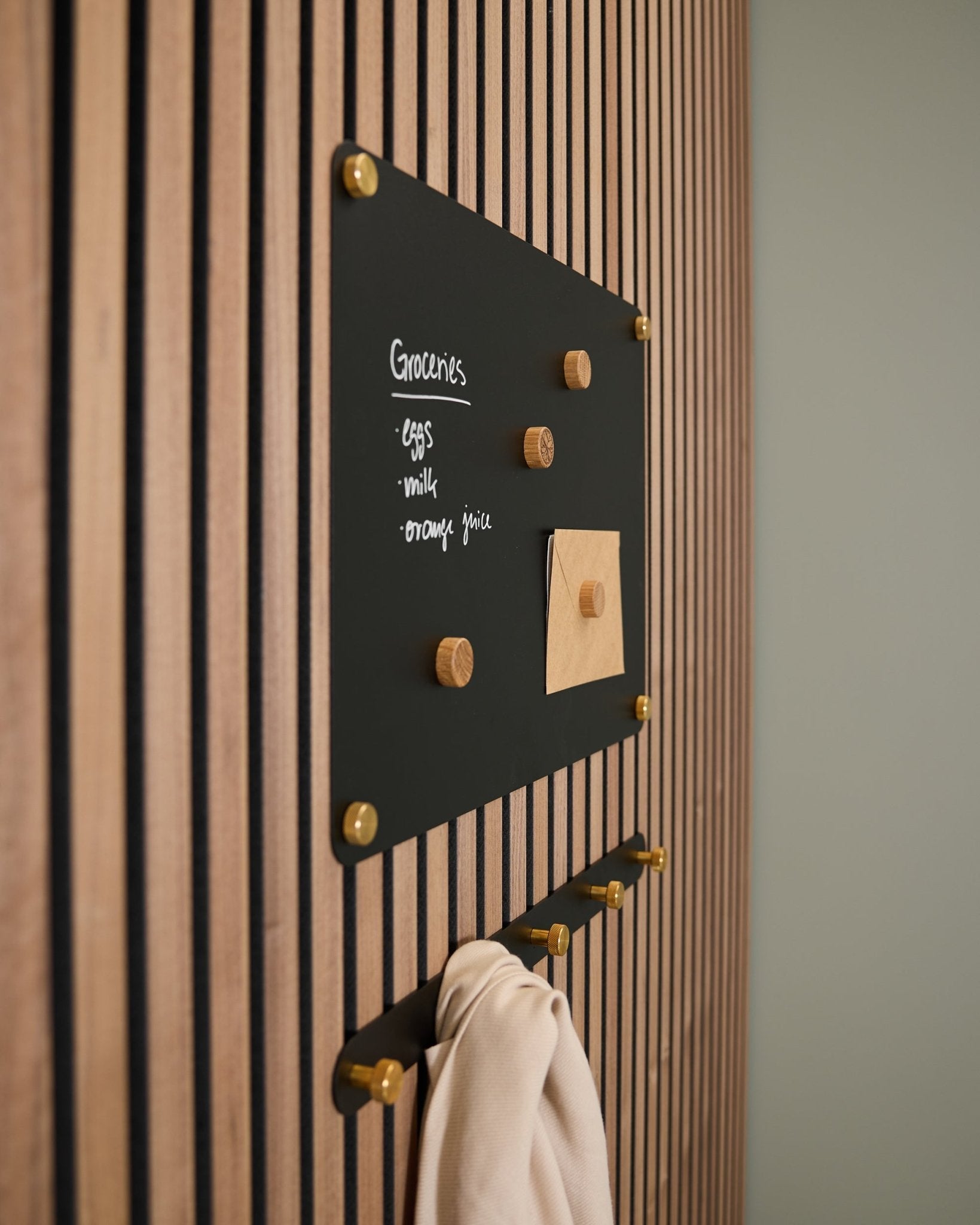 Een small black Norr Magnetic Board van WoodUpp, gemonteerd op een Akupanel akoestisch houten paneel, toont een boodschappenlijst en notitie, met daaronder haken waaraan een lichte doek hangt.