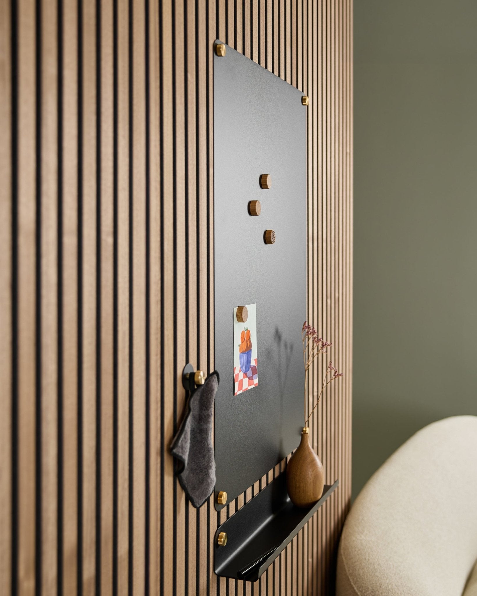 Het akoestische magnetische bord van WoodUpp Norr large black met houten magneten, prikbord, plank en vaas is aan de muur gemonteerd naast een beige stoel.