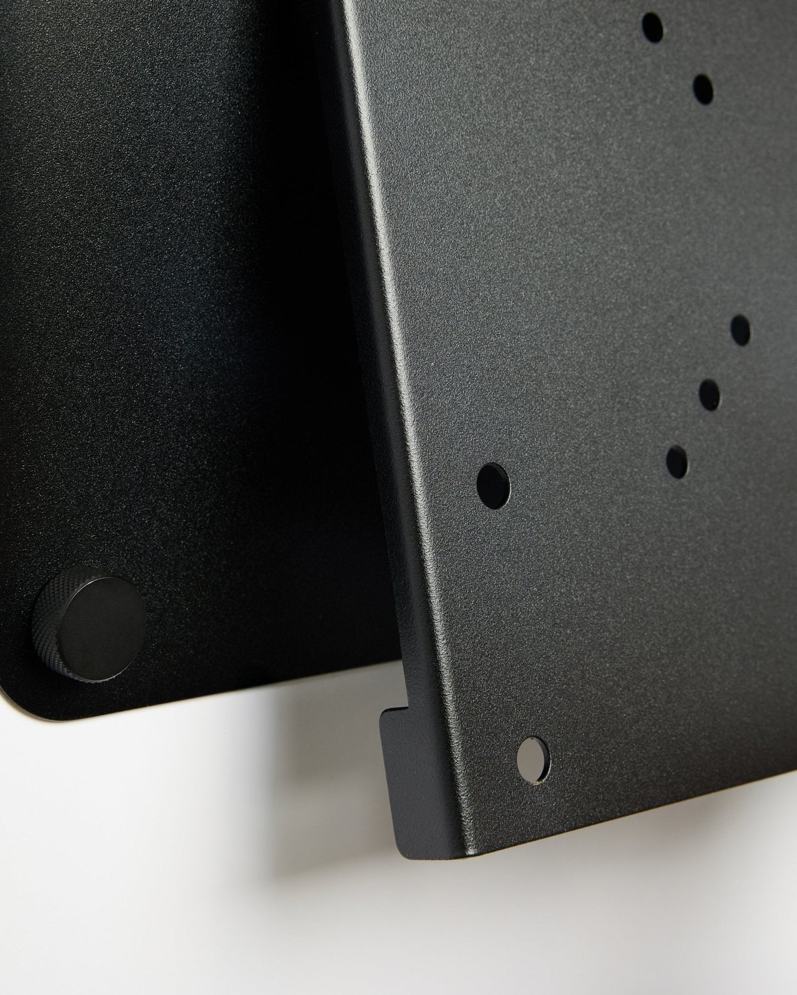 Close-up van de Fjell TV Mount (small) van WoodUpp, een black metalen beugel met ronde openingen en een verstelknop, weergegeven tegen een witte achtergrond.