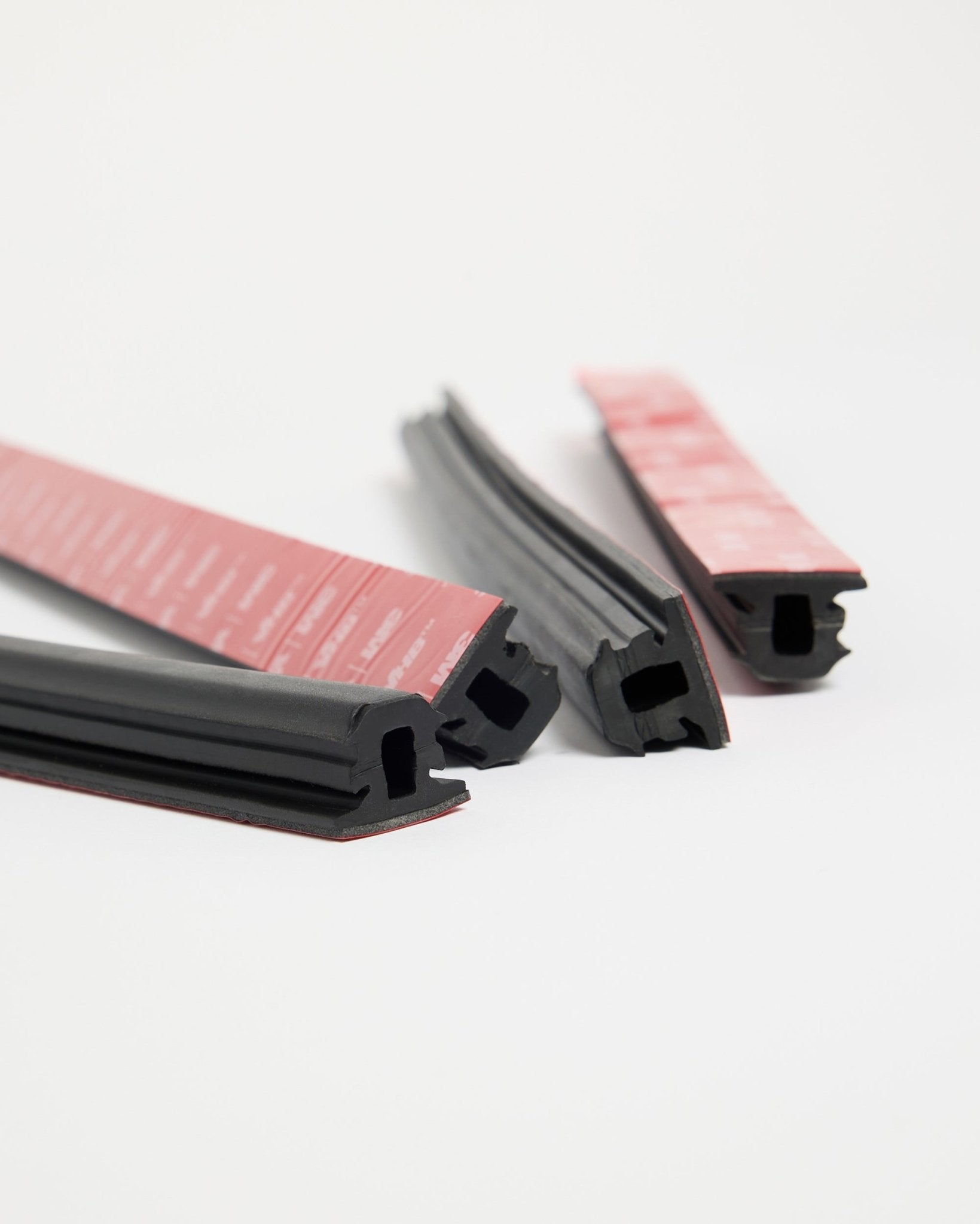 Vier WoodUpp Dupps Pop-on Profile black rubberstrips met rode kleeflaag, ontworpen voor Akupanel installatie, in verschillende richtingen neergelegd op een wit oppervlak.