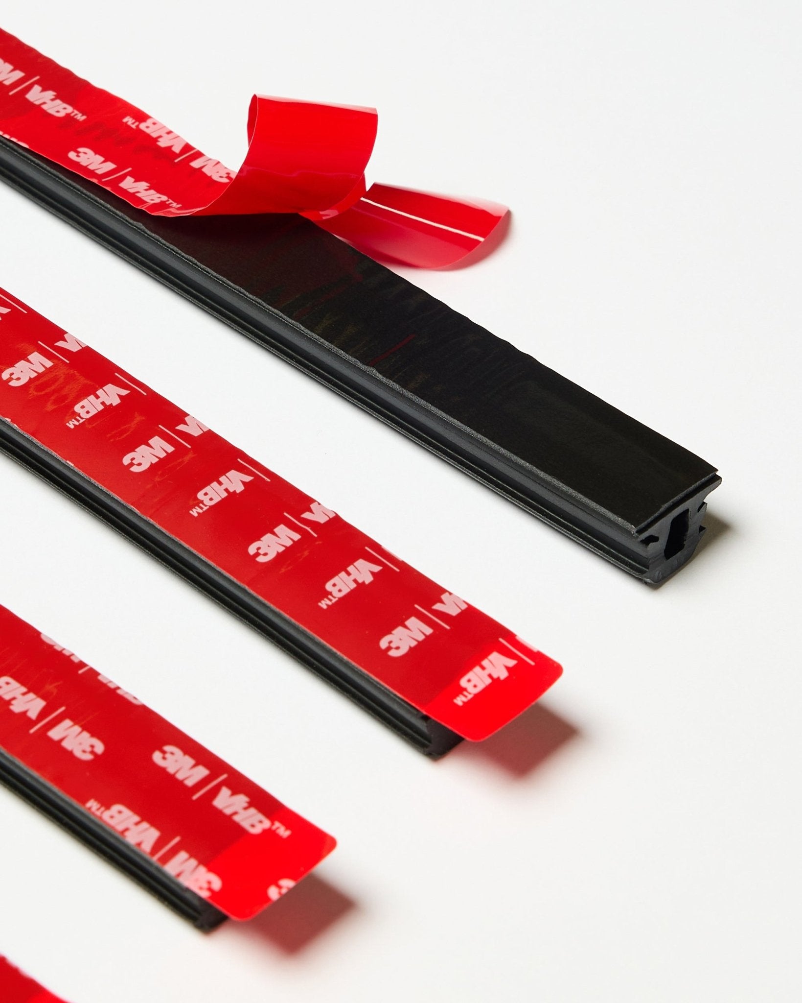 Set van vier black WoodUpp CREATE Pop-on Profile strips met zichtbare rode 3M-kleeflaag, waarvan één strip deels is losgehaald, gepresenteerd op een wit oppervlak.