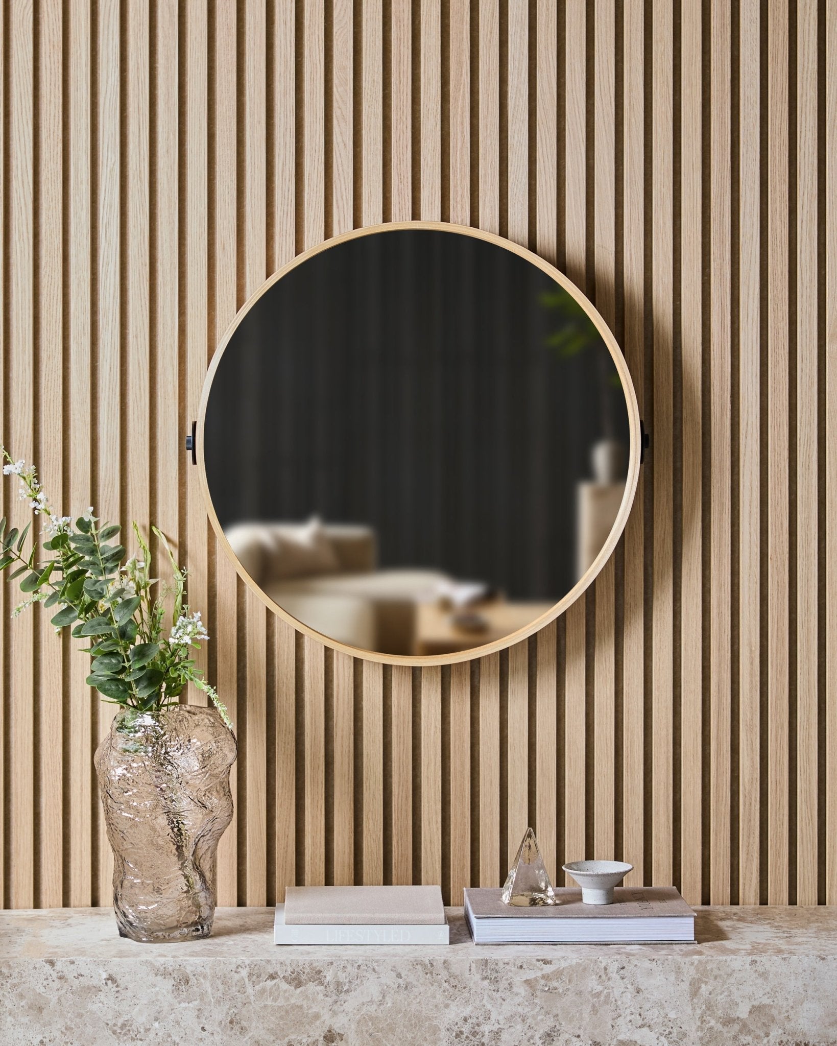 De Linn | Mirror in Natural Oak van WoodUpp is gemonteerd op een Nordic lattenwand Akoestisch paneel boven een stenen plank met een glazen vaas, boeken en decoratieve items.