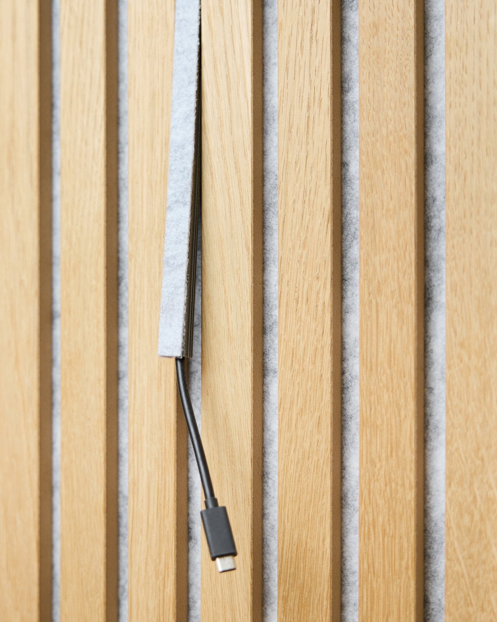 Een black USB-C-kabel loopt door een grey WoodUpp Dupps Pop-on Cable Cover tussen vertical houten akoestische latten met grey vilten achterkant.