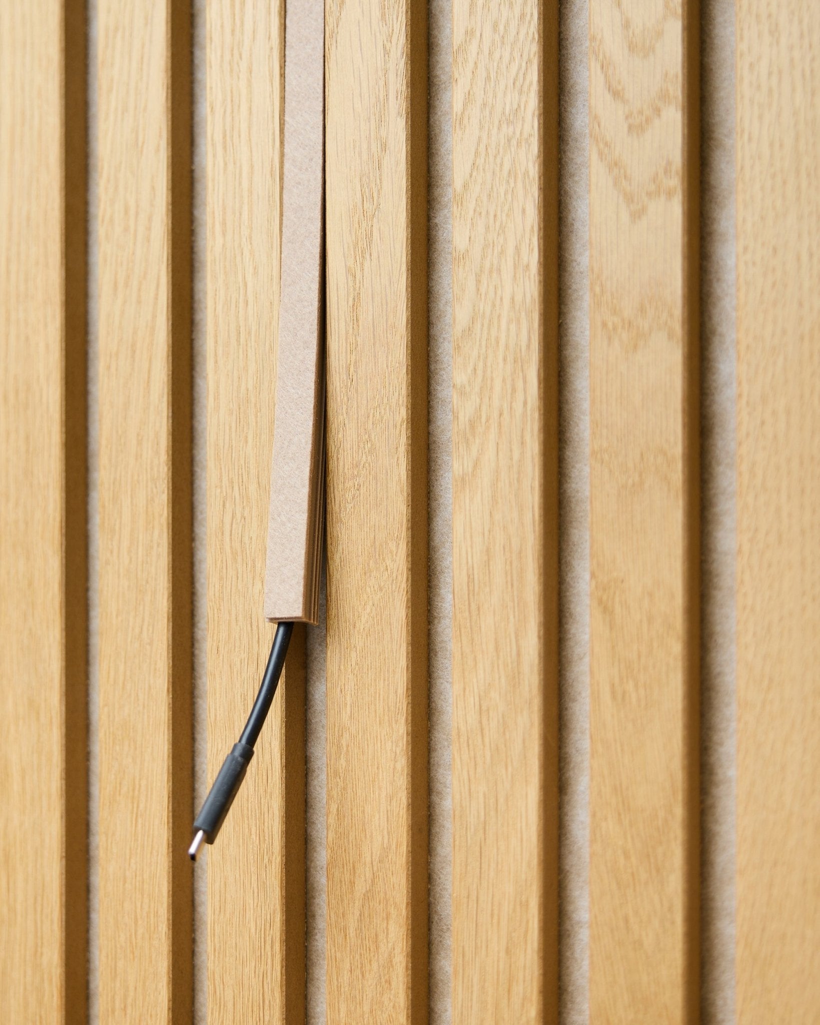 Een black kabel met kabelbeheer is deels weggewerkt in een WoodUpp Dupps Beige Pop-on Cable Cover, gemonteerd op vertical Akupanel akoestische houten latten.