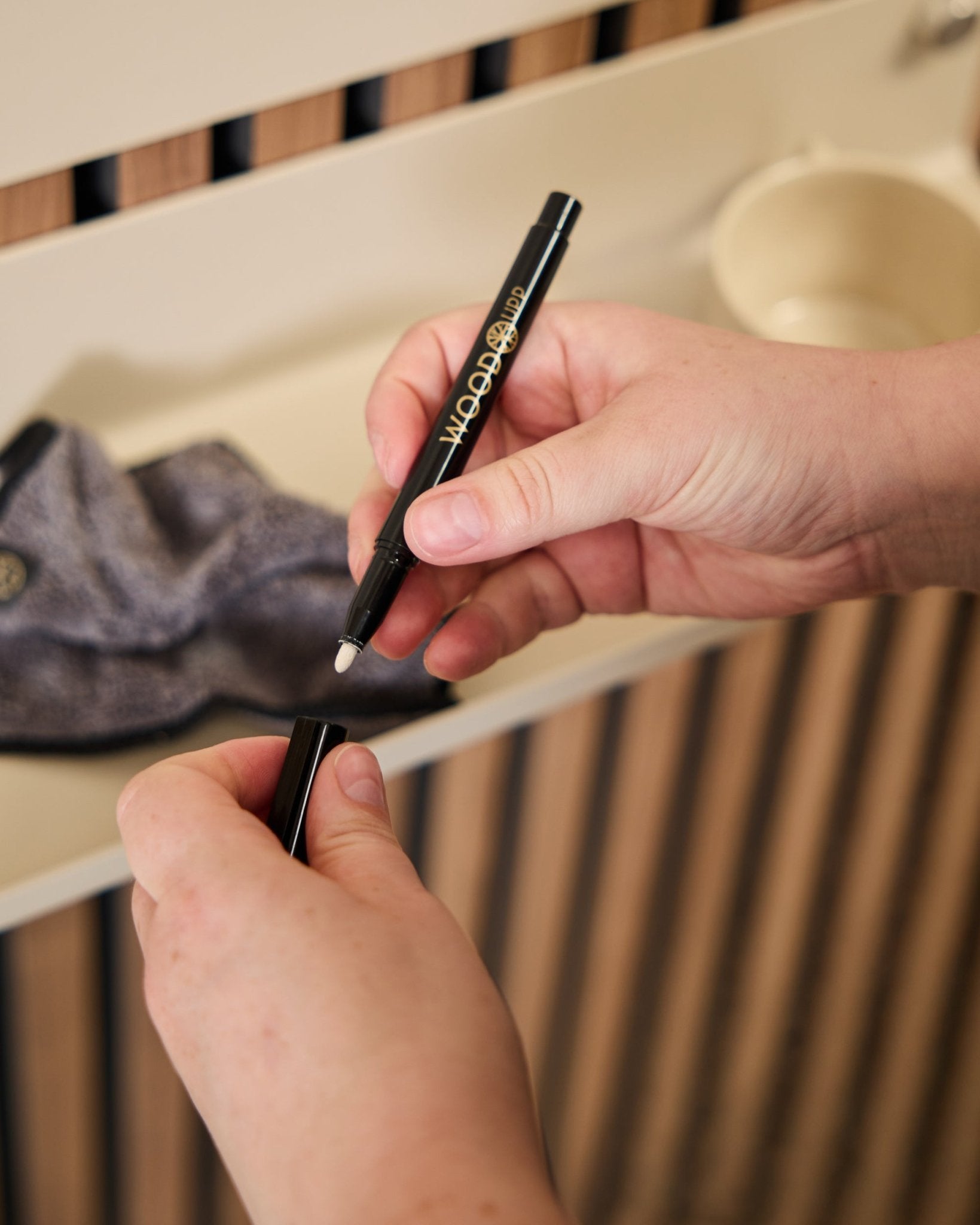 Iemand houdt een open WoodUpp Norr | Blackboard set marker vast naast een microvezeldoek en een beige beker, producten die zijn ontworpen voor gebruik met akoestische houten panelen.