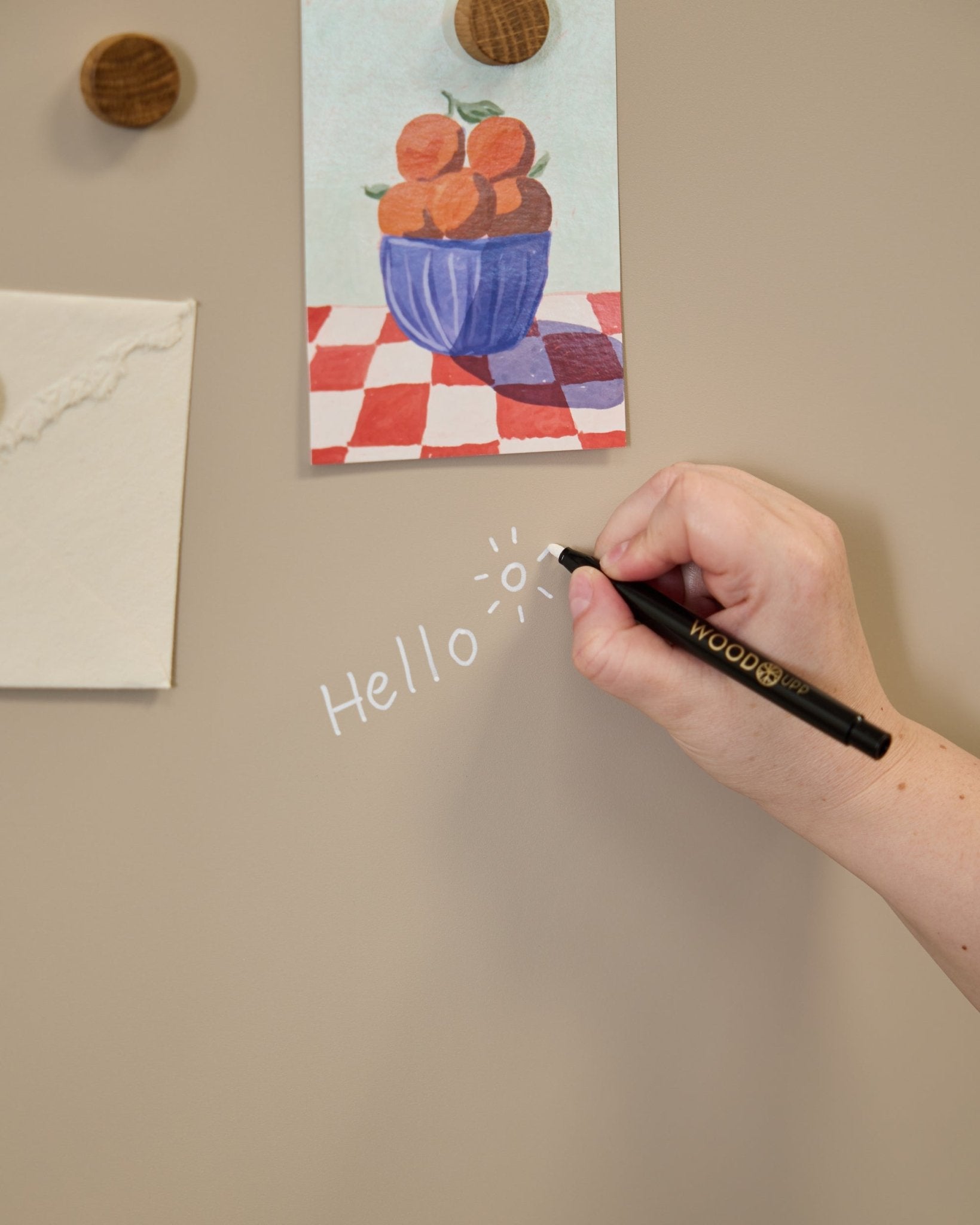 Een hand schrijft hallo met een zonnetje op het WoodUpp Norr Blackboard, naast kunst die met ronde magneten is bevestigd op een Akoestisch paneel.
