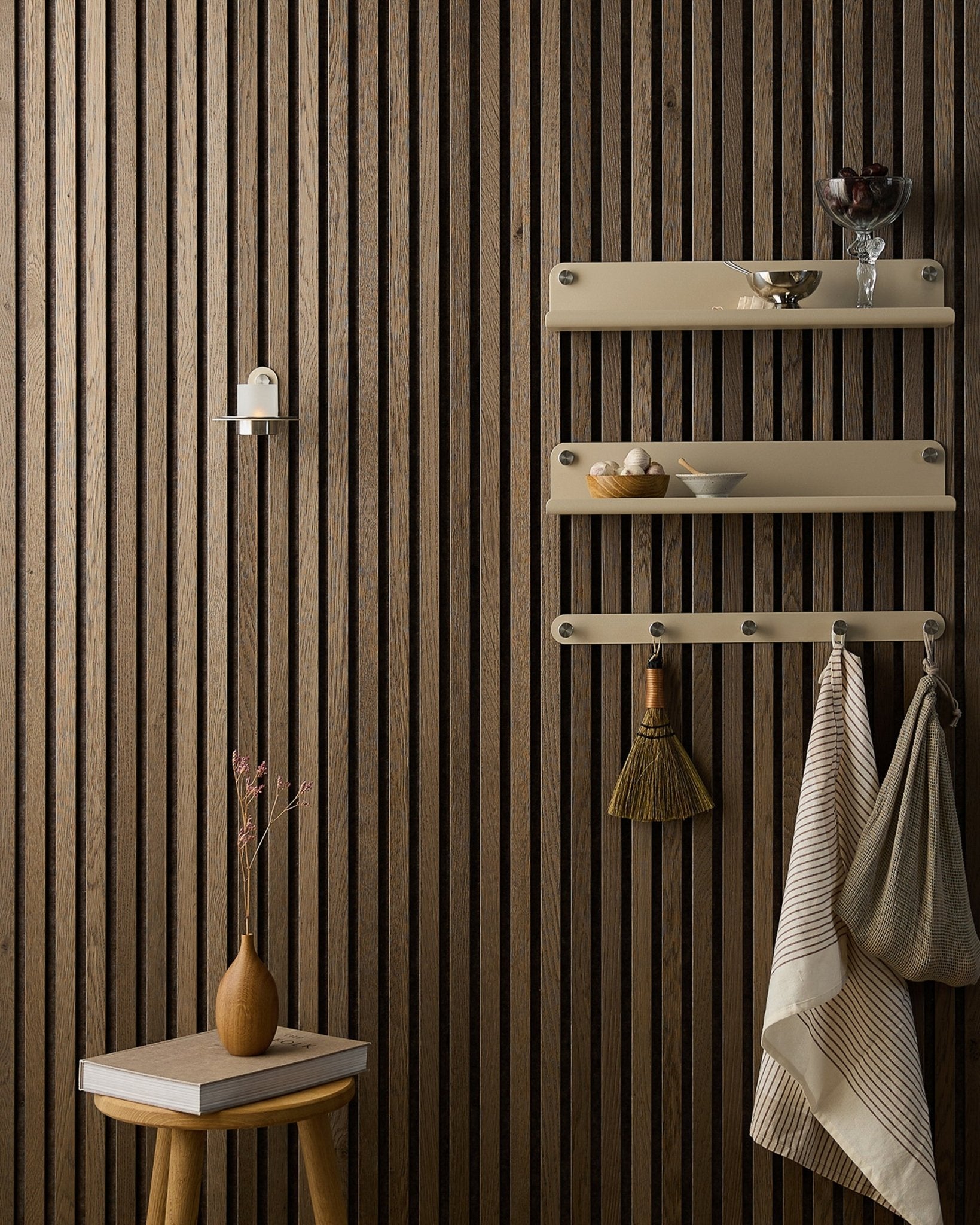 Een houten kruk met droogbloemen staat naast een WoodUpp Akupanel Akoestisch paneel, naast beige planken met servies, een borstel, handdoeken en een Norr Candle Holder.