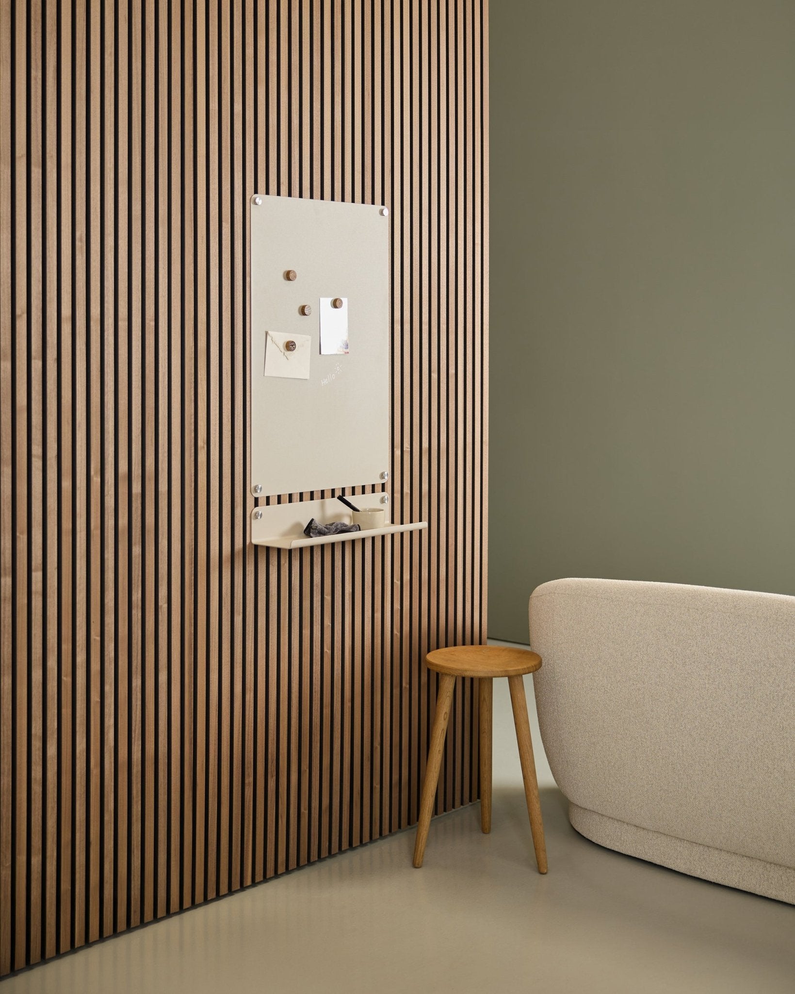 Minimalistisch interieur met beige bank en houten kruk, met een large beige WoodUpp Norr magneetbord gemonteerd op een Nordic Akupanel akoestisch houten paneel bij een groene muur.