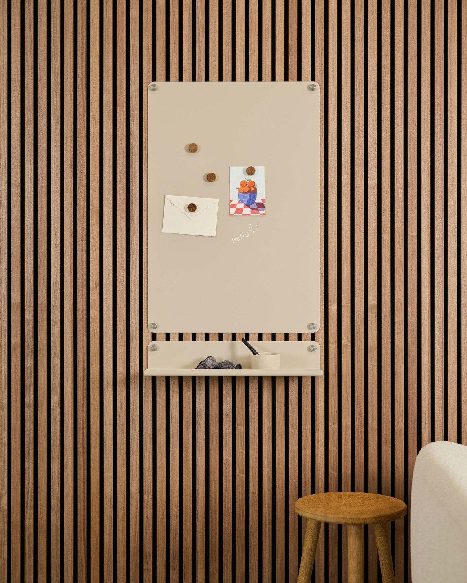 Het Beige Norr magneetbord van WoodUpp is gemonteerd boven een small plank aan een wand met Akupanel akoestische houten panelen, met een kruk en witte bank in de buurt.