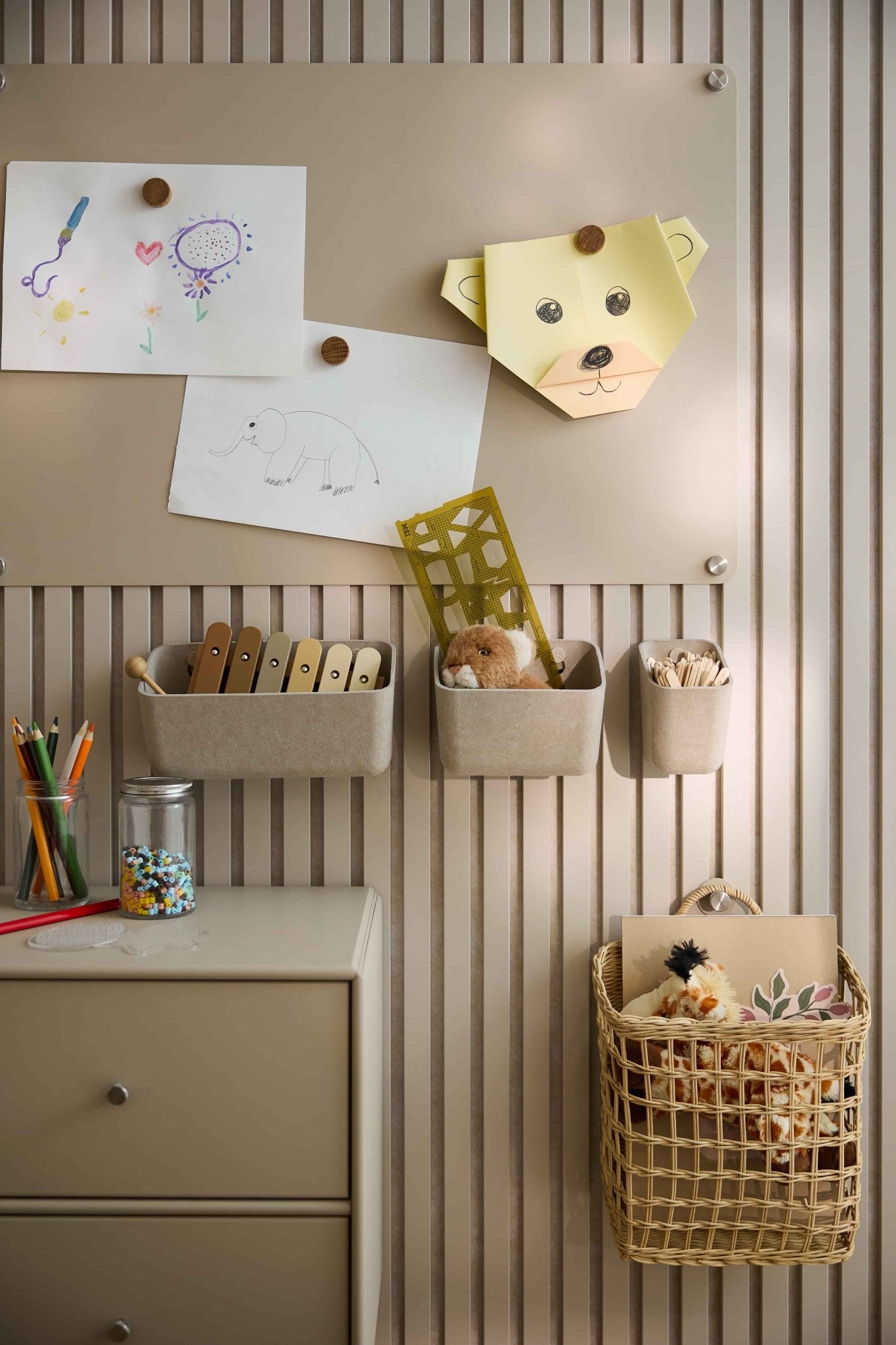 Op een beige muur met een WoodUpp-akoestisch paneel zijn kinderkunstwerkjes en speelgoed uitgestald, waaronder vilten bakjes, mandjes, een lade en een perforatieplaat.