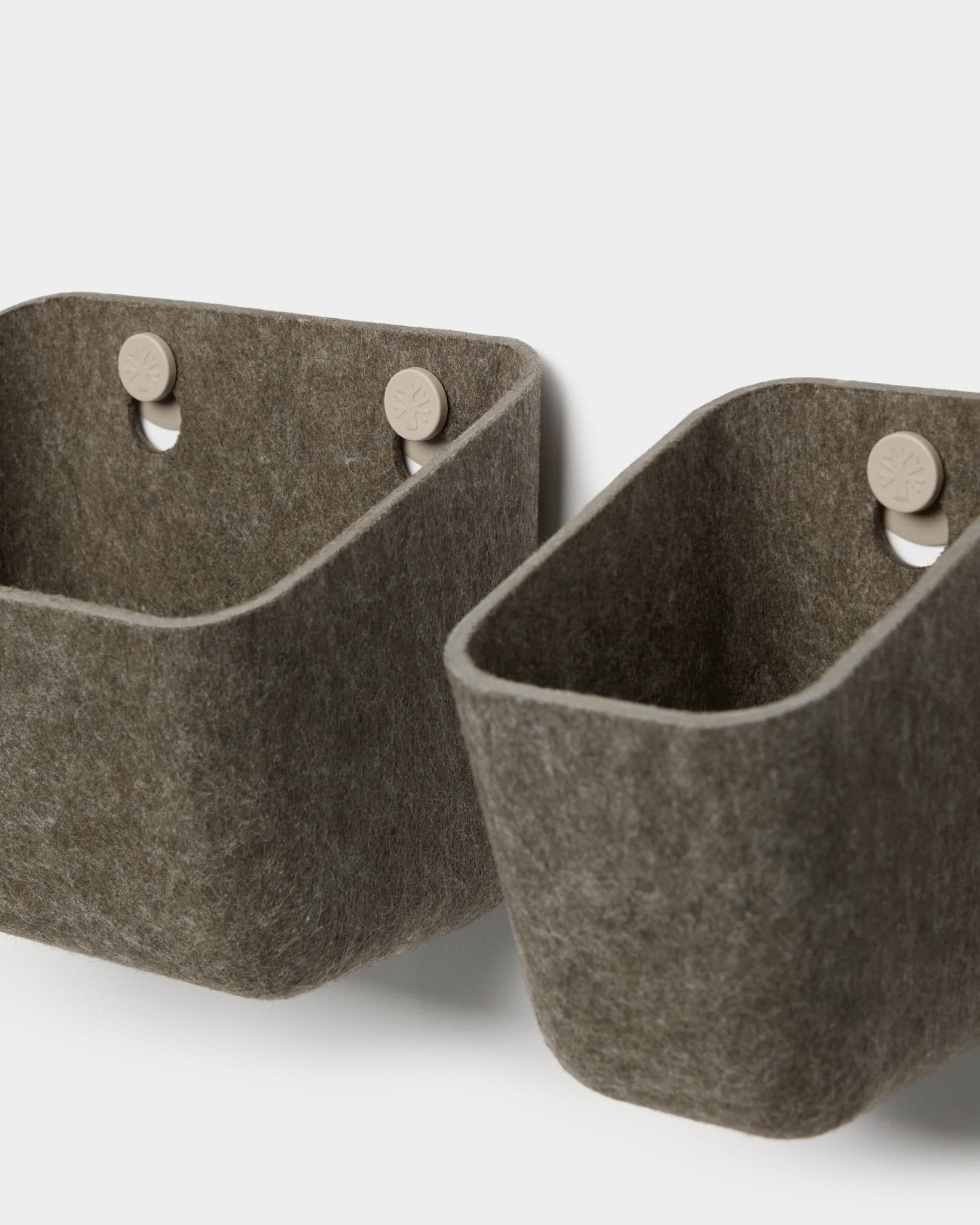 Twee Luva-viltbakjes in donker, warm grijs van WoodUpp hangen aan beige haken die aan een akoestisch paneel zijn bevestigd, en zorgen zo voor minimalistische wandopbergruimte met afgeronde hoeken.