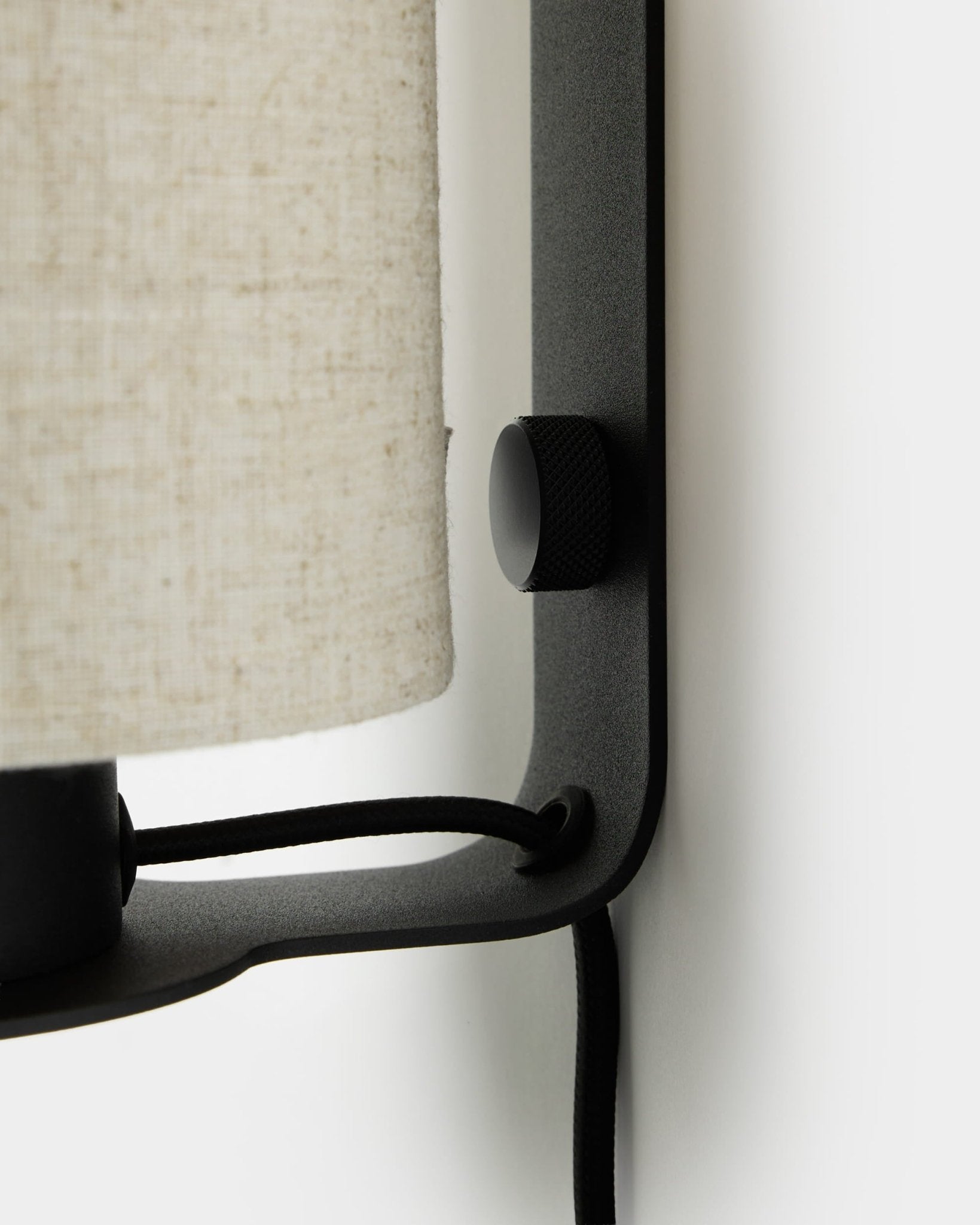 Close-up van de WoodUpp Feya wandlamp in black en beige, met een knop met textuur, een kap van linnen-katoen en een zichtbaar snoer, gemonteerd op een witte muur.