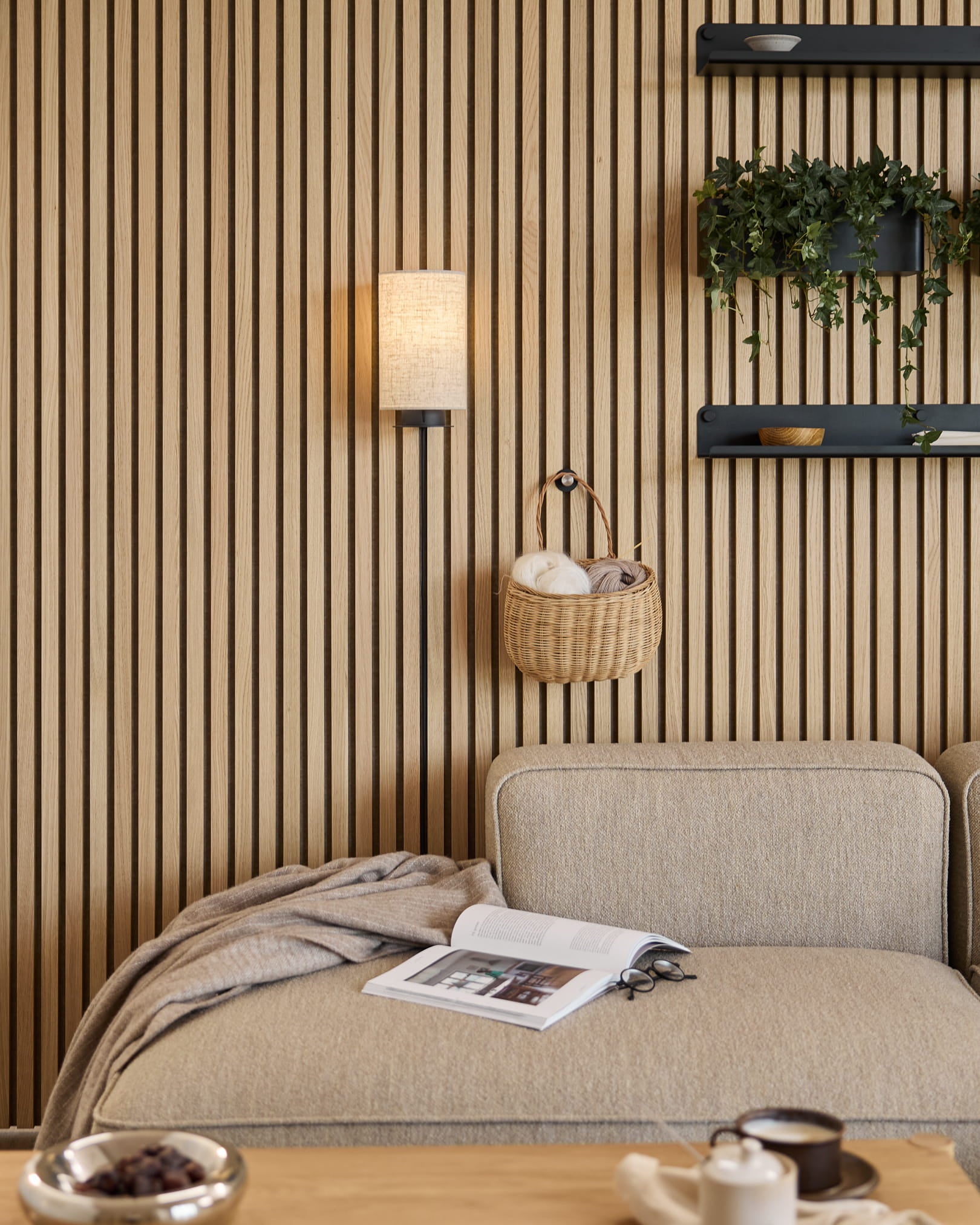 Bank in Beige met plaid en boek naast een WoodUpp Akupanel akoestische lattenwand, planken met planten en decoratie, en een Feya wandlamp in Black/Beige.