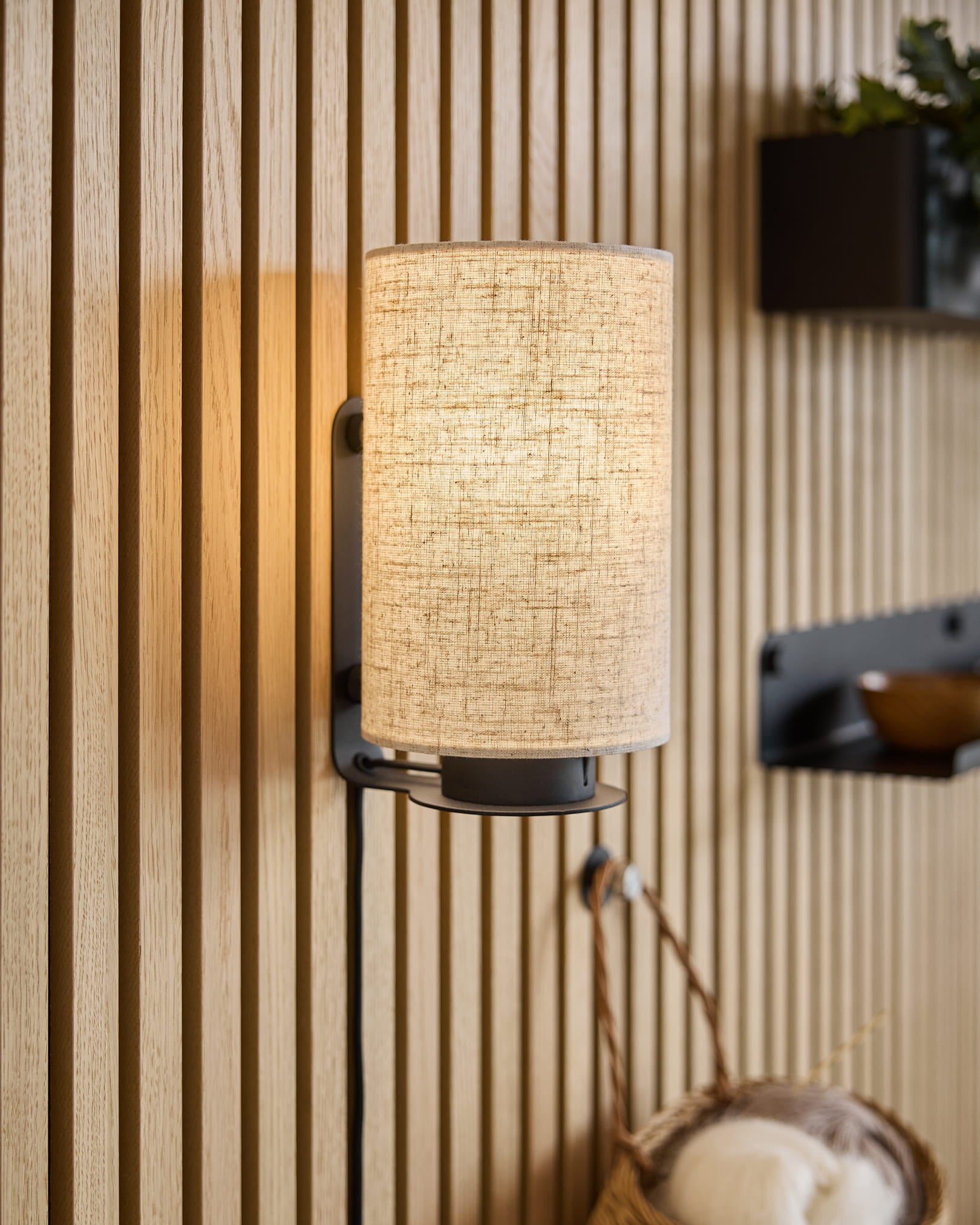 Een moderne wandarmatuur met een stoffen kap werpt warm licht op Akupanel akoestische houten panelen, aangevuld met minimalistische planken en een geweven mand.