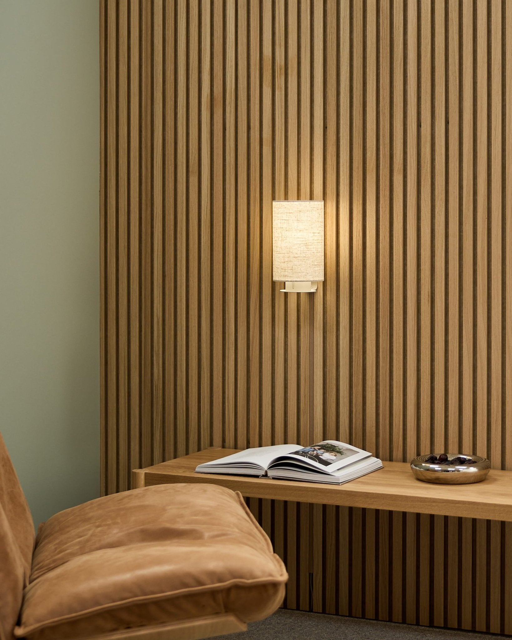 Minimalistische Scandinavische ruimte met een tan-kleurige stoel, WoodUpp Akupanel akoestische lattenwand, Feya wandlamp in Beige/Beige, een open boek op een bankje en een kom.