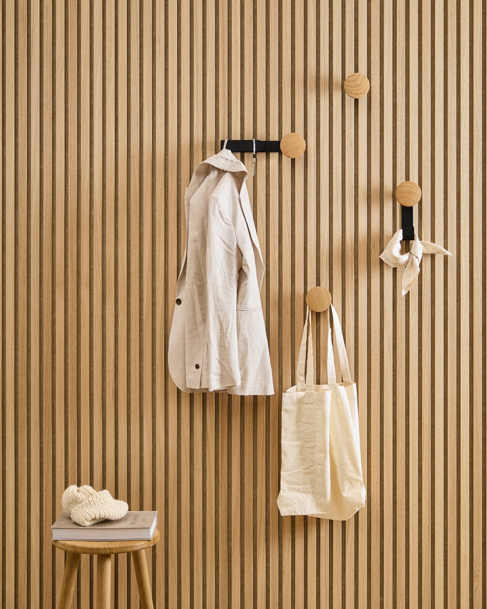 Een beige jas, tas en sjaal hangen aan WoodUpp’s Linn Horizontal Hanger in Natural Oak; op een krukje ernaast liggen een boek en een gebreid item.