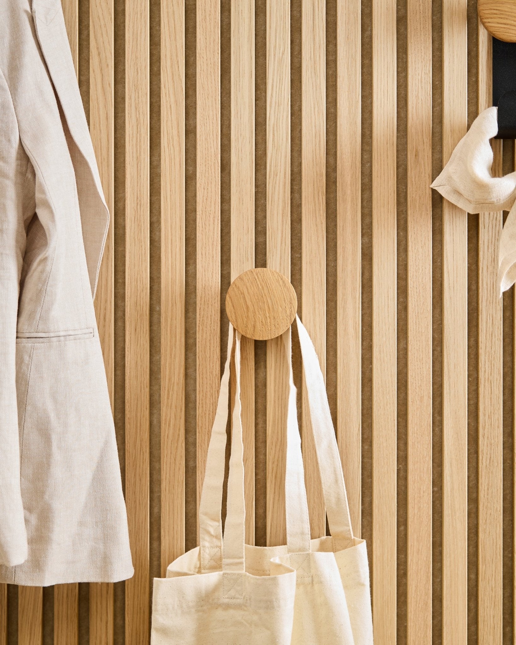 Een beige jas en tote bag hangen aan de Linn I Single Hanger in Natural Oak van WoodUpp, gemonteerd op een vertical Akupanel akoestisch houten lattenpaneel.