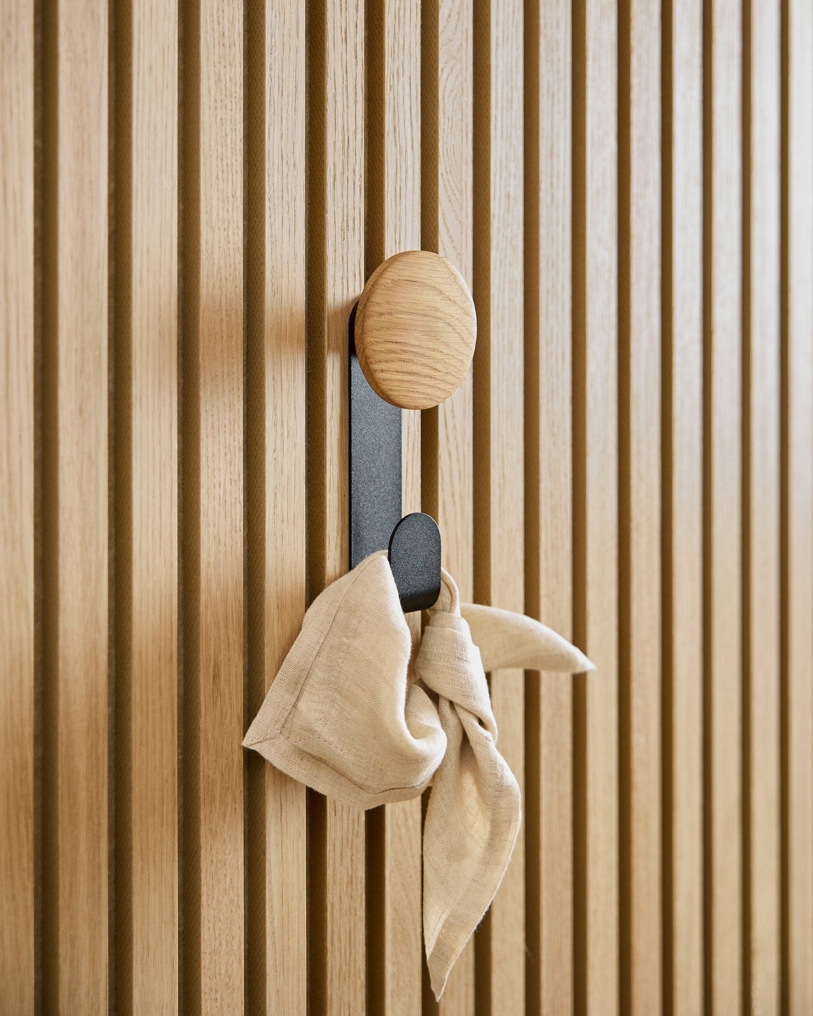 Een doek van beige hangt aan de Linn Vertical Hanger in Natural Oak van WoodUpp, gemonteerd op een akoestisch lattenwandpaneel van vertical Akupanel.