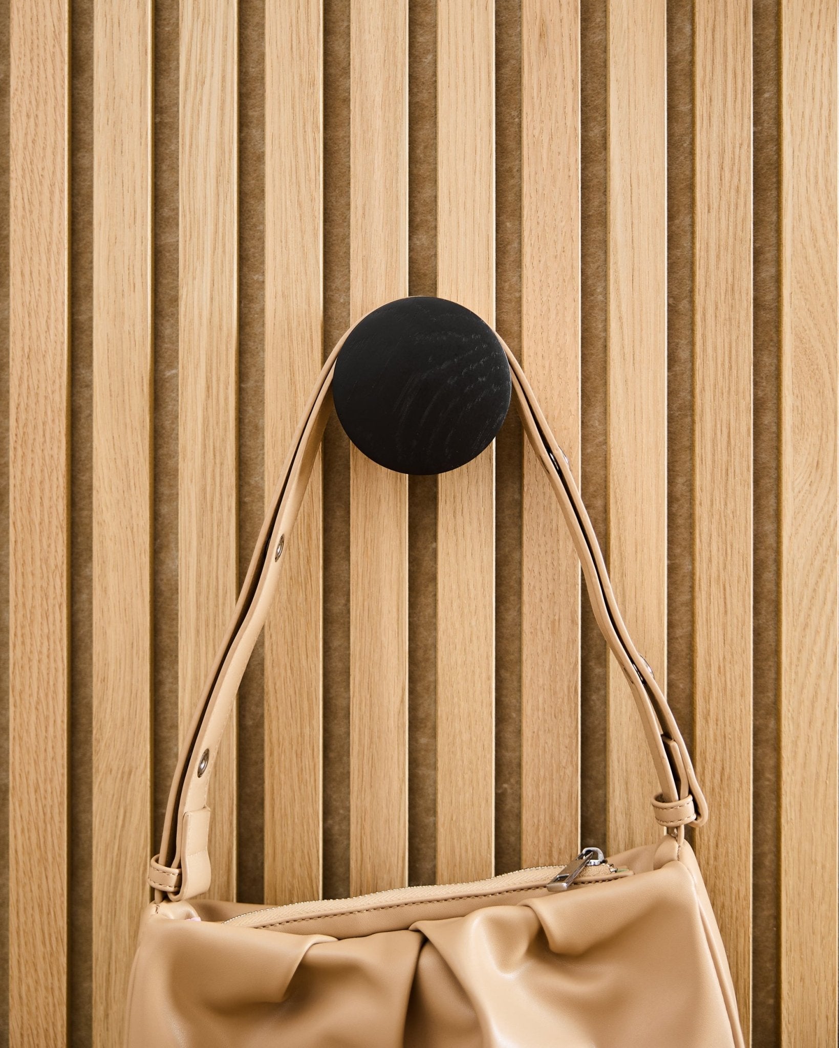 Beige handtas hangend aan een WoodUpp Linn | Single Hanger in Black Oak, gemonteerd op een licht houten Akupanel akoestisch lattenwandpaneel.