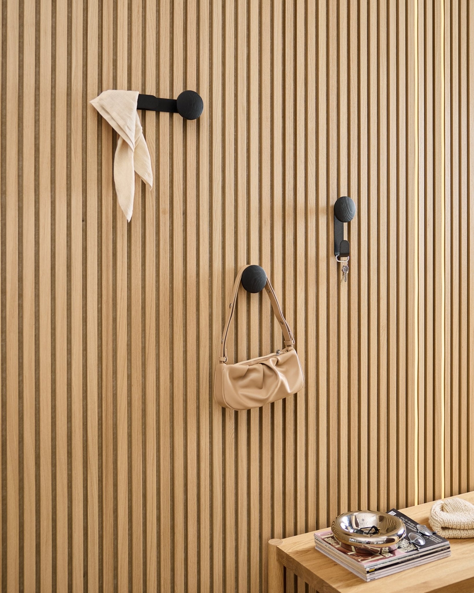 WoodUpp Linn Single Hanger in Black Oak draagt een sjaal, beige tas en sleutels op een black Akupanel akoestisch houten paneel; bankje met boeken en metalen schaal ernaast.