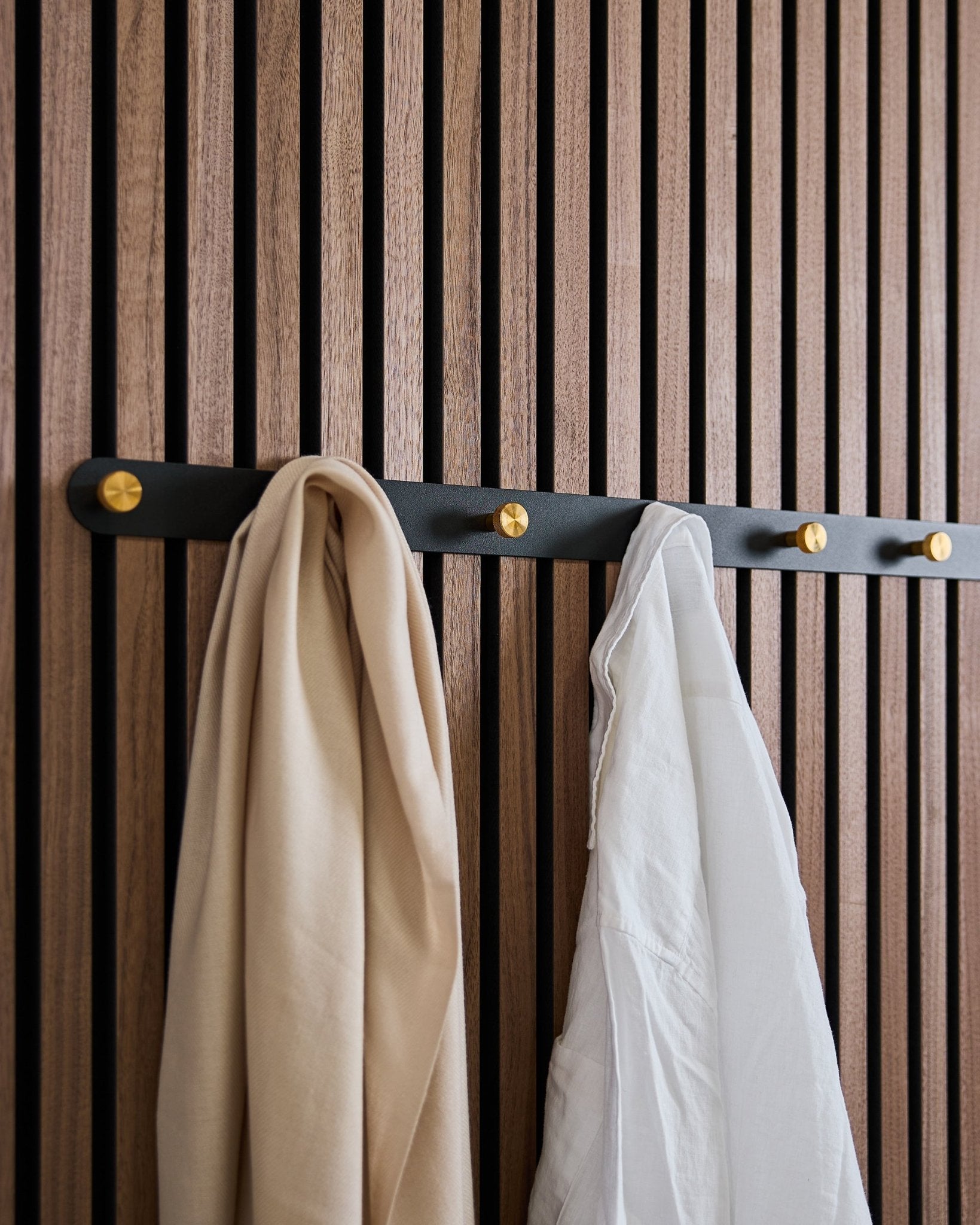 De WoodUpp Norr kapstok in black/brass (X7) met een beige sjaal en wit overhemd hangt aan een akoestisch lattenwandpaneel van hout in Scandinavische stijl.